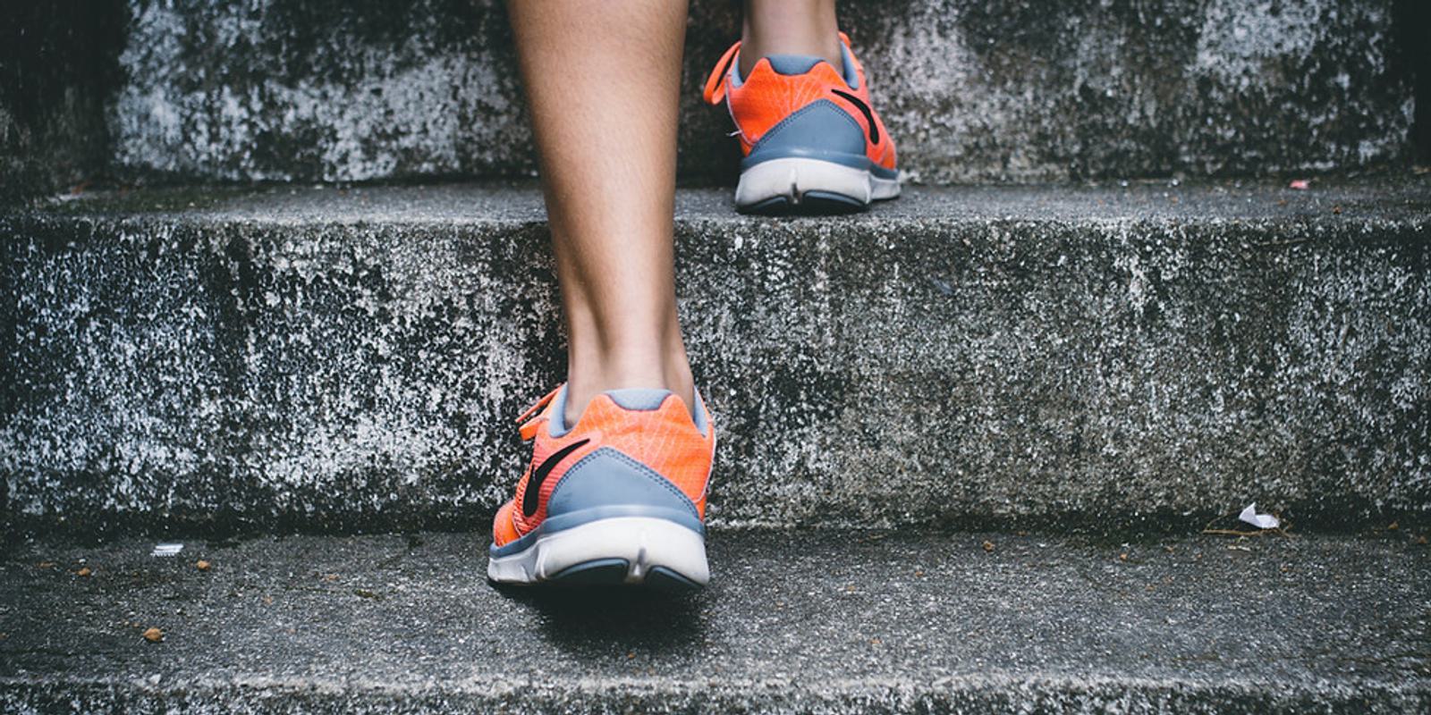 Close-up of a person's lower legs and feet wearing bright orange and gray athletic shoes, walking up outdoor concrete stairs. The texture of the rough, weathered stairs contrasts with the vibrant colors of the shoes.