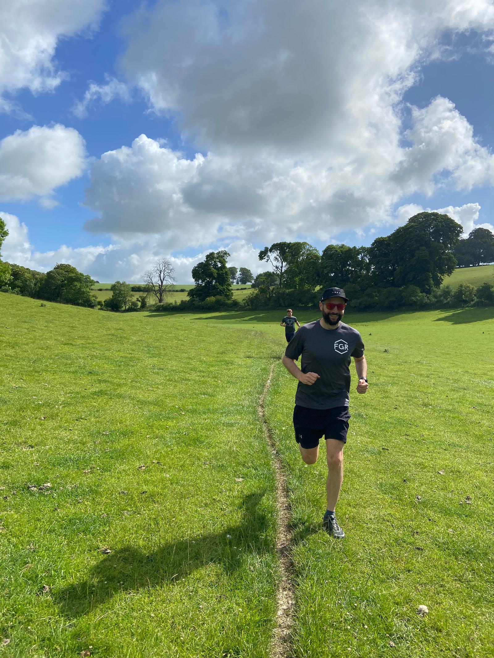 A man in a black shirt, shorts, sunglasses, and a cap is running in a grassy field with a clear path under a bright, partly cloudy sky. Another person is running behind him. Trees and hills are in the background.