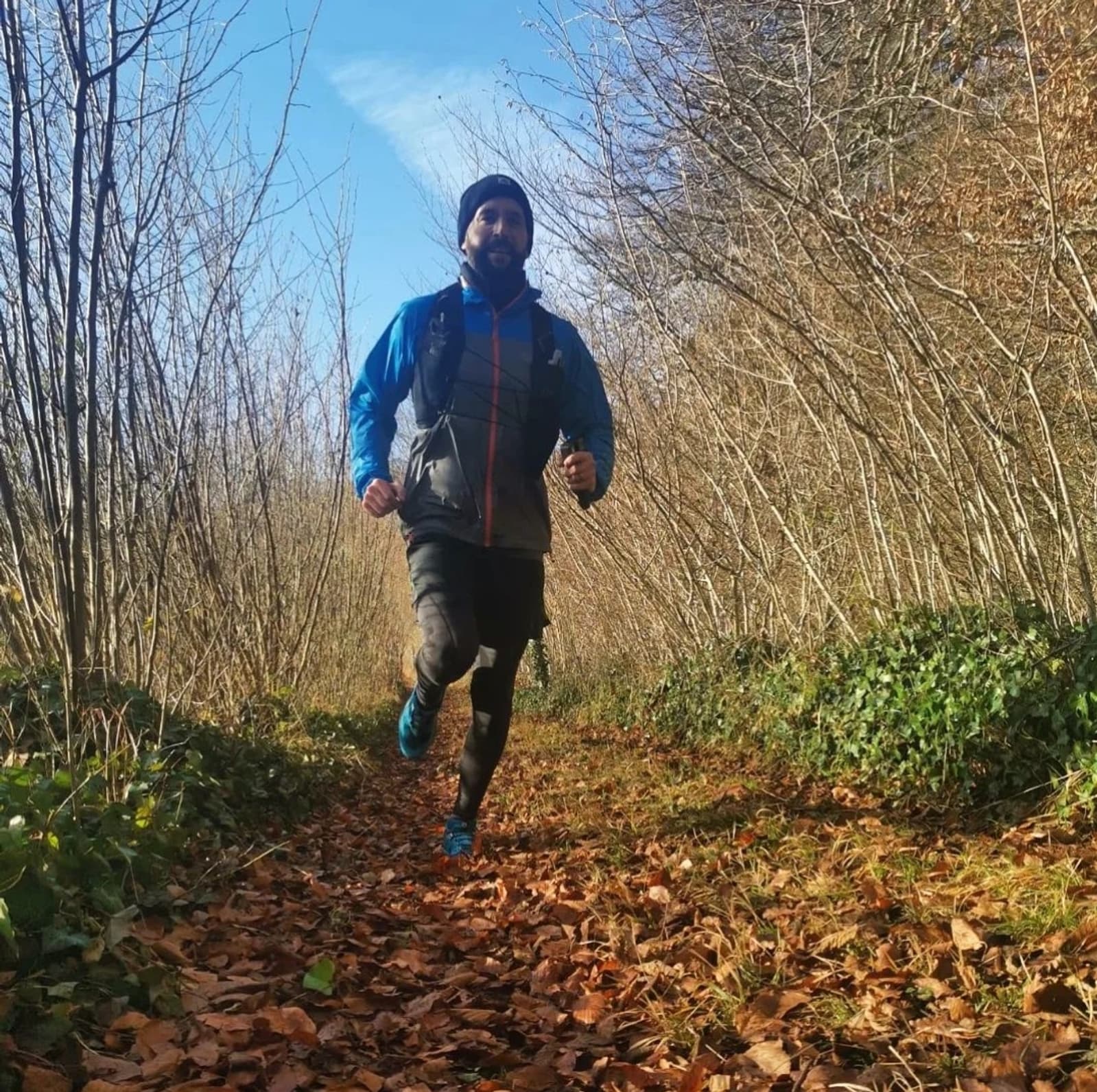 A person is trail running through a forest path covered in fallen leaves. They are wearing a black beanie, a gray and blue jacket, and black leggings. Bare trees and a blue sky are in the background.
