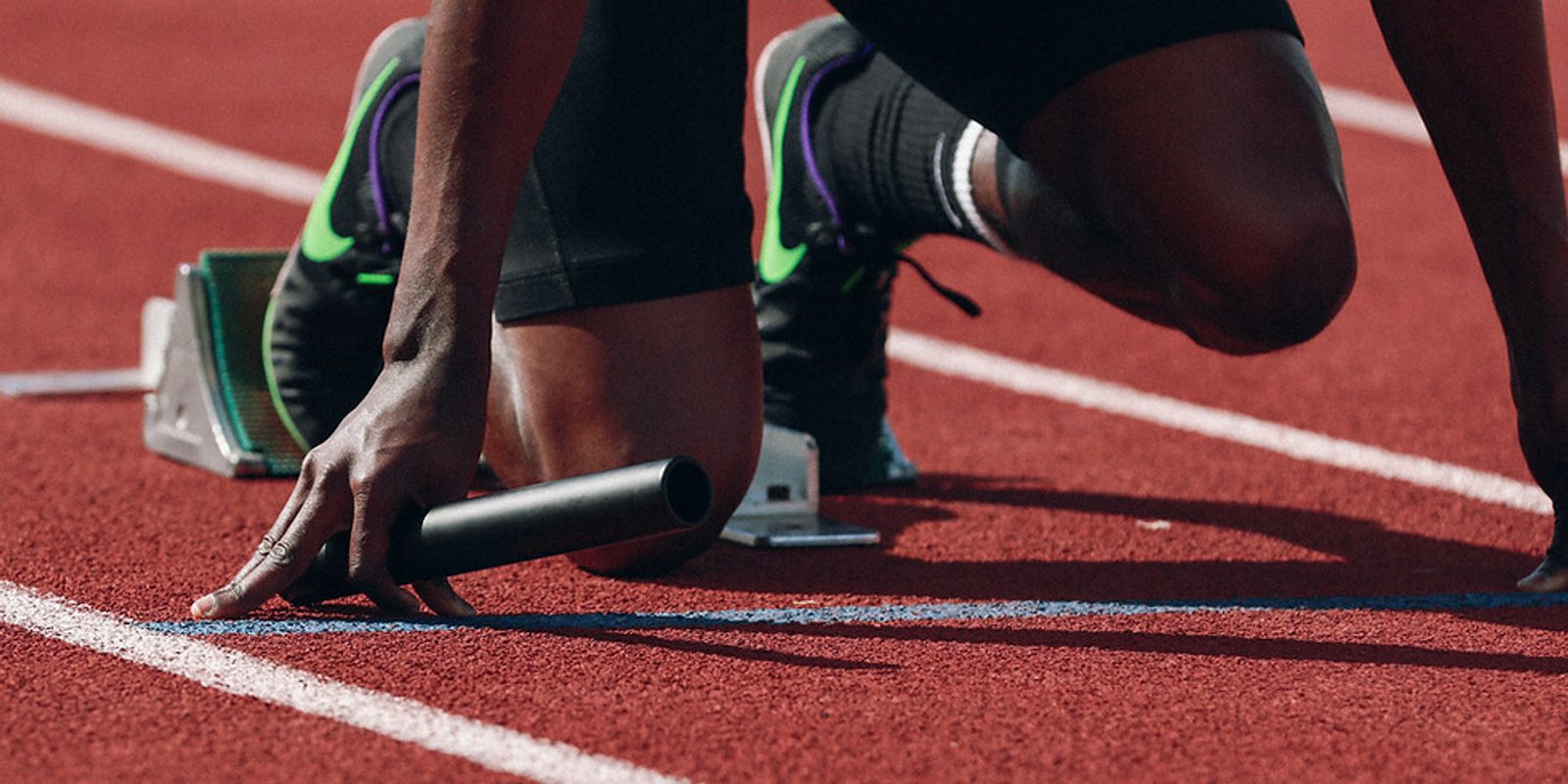 A sprinter crouches in the starting blocks on a red running track, holding a baton, preparing for a relay race. The athlete's feet are secured, and they are wearing black athletic shoes with green accents.