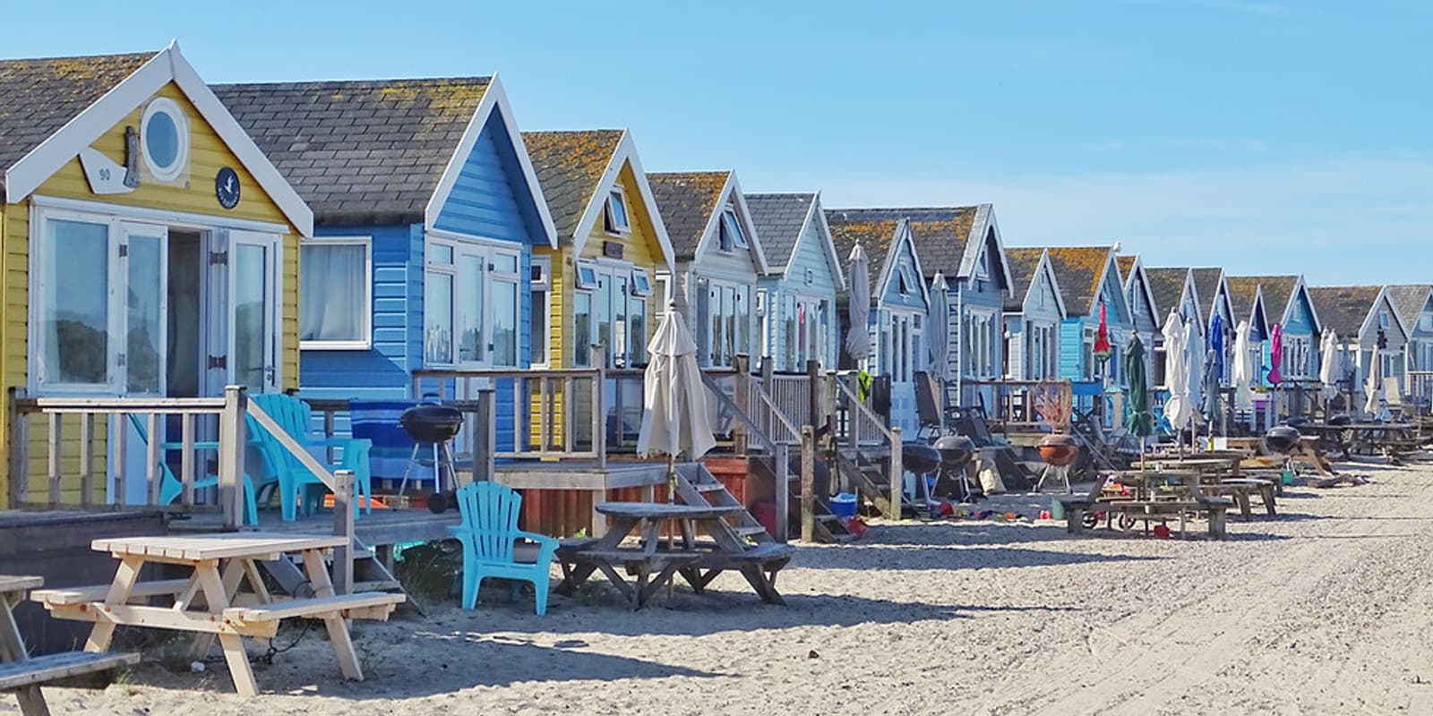 Seaside view of colorful beach huts lined up along a sandy shore. Each hut has a small porch, some with outdoor furniture and umbrellas. Picnic tables and scattered items are visible on the beach, and the sky is clear and blue.
