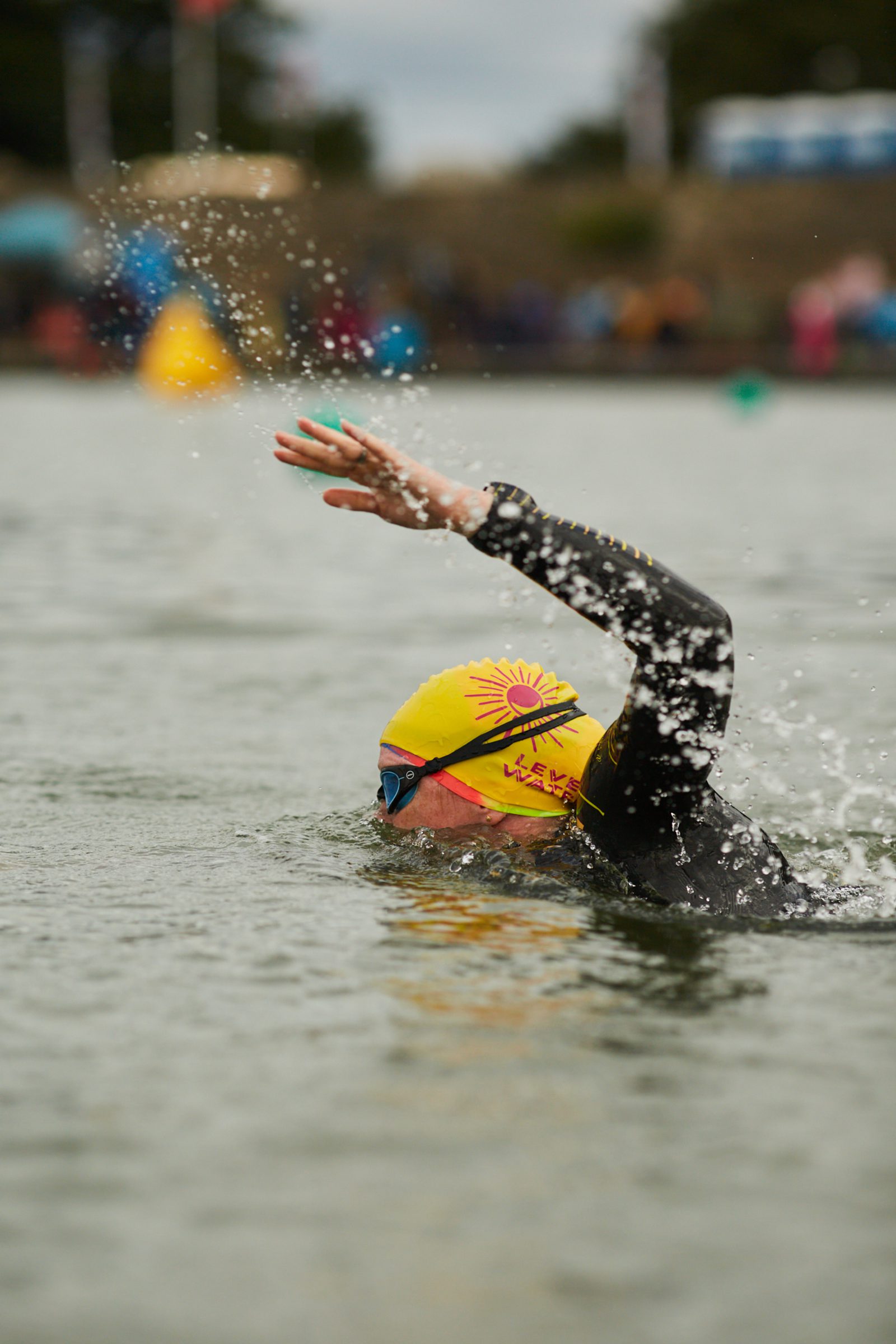 A swimmer wearing a yellow swim cap with a sun design, blue goggles, and a black wetsuit swims in open water. Their left arm is outstretched, splashing water droplets, with a blurred background of spectators and colorful buoys.