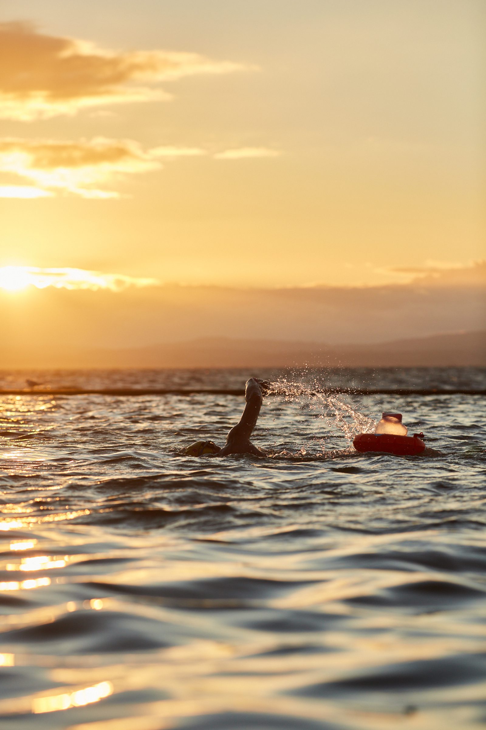A person swims in the ocean at sunset, with one arm raised out of the water. A red float or buoy is visible next to them. The sky is bathed in warm orange and yellow hues as the sun sets on the horizon. Nearby water ripples with gentle waves.