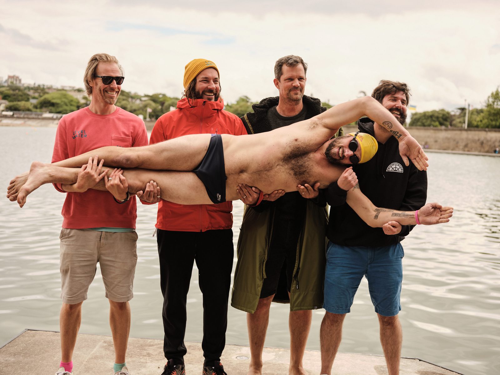 Five men pose in front of a body of water. Four of them stand while one man, wearing a yellow swim cap and black swimming briefs, is being held horizontally by the others. They are all smiling and appear to be enjoying the moment.