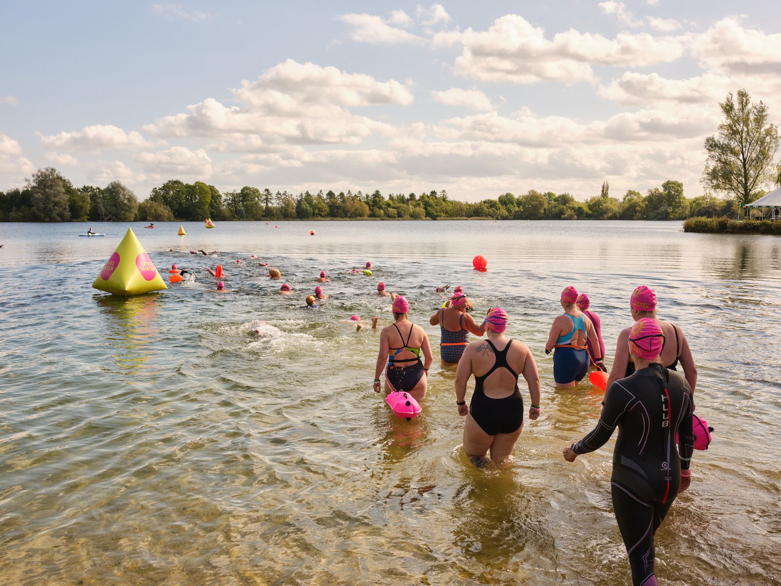A group of swimmers in wetsuits and pink swim caps enter a lake for a race. Several swimmers are already in the water, heading towards brightly colored buoys. The lake is surrounded by trees, and the sky is partly cloudy with ample sunshine.