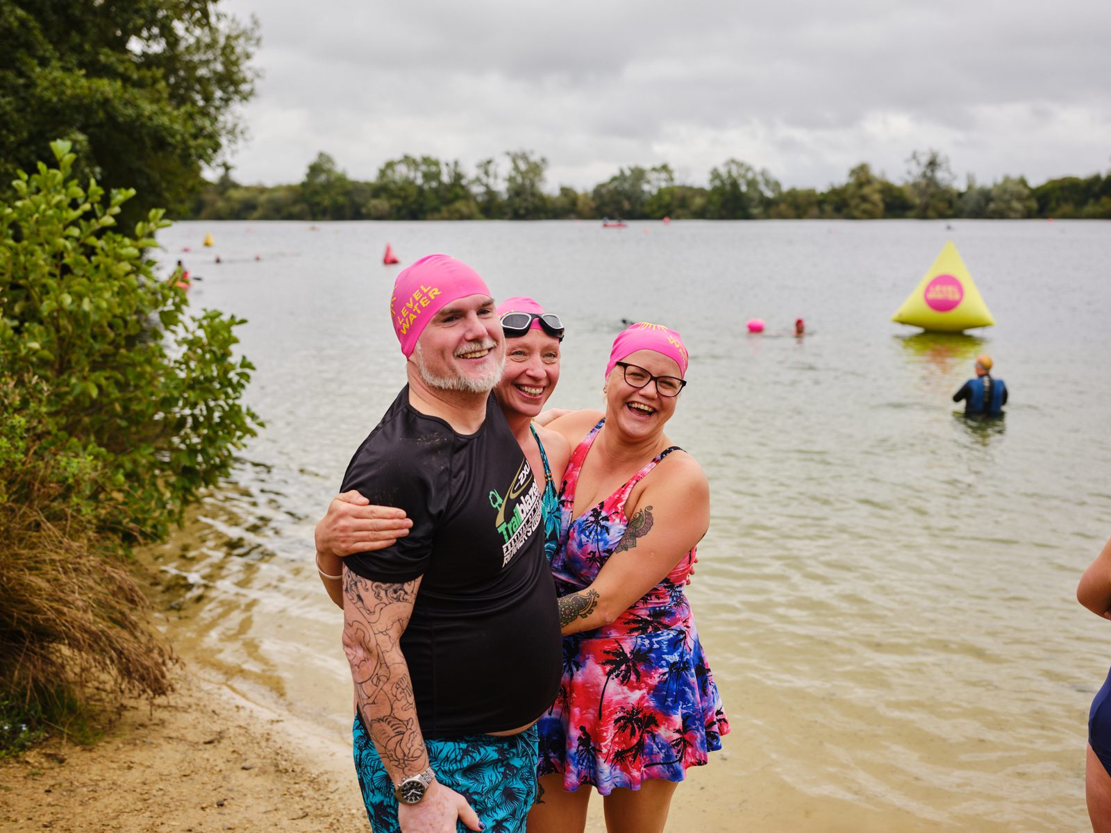Three smiling individuals in colorful swimwear and pink swim caps, standing at the edge of a lake. The person on the left in a black shirt has tattoos. The person in the middle is holding the person on the right, who is in a floral swimsuit. Swimmers and buoys are visible in the water.