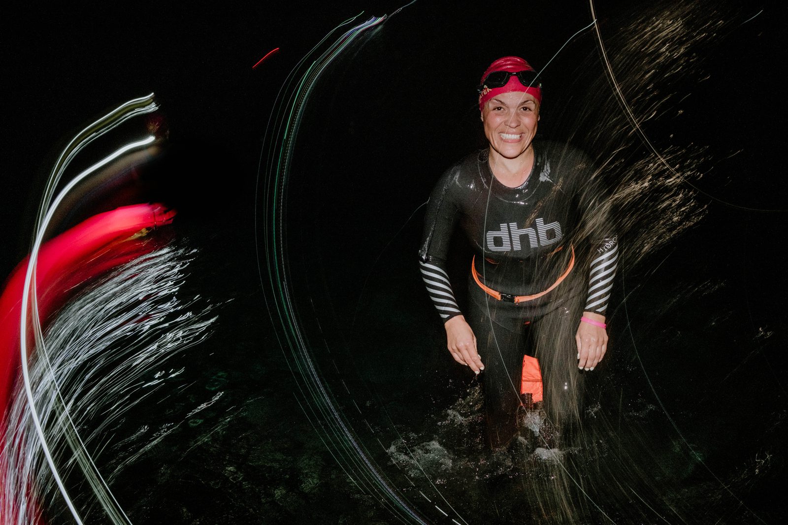A person wearing a black wetsuit, pink swim cap, and goggles is smiling while standing in water at night. Light trails surround them, adding a dynamic effect to the scene.