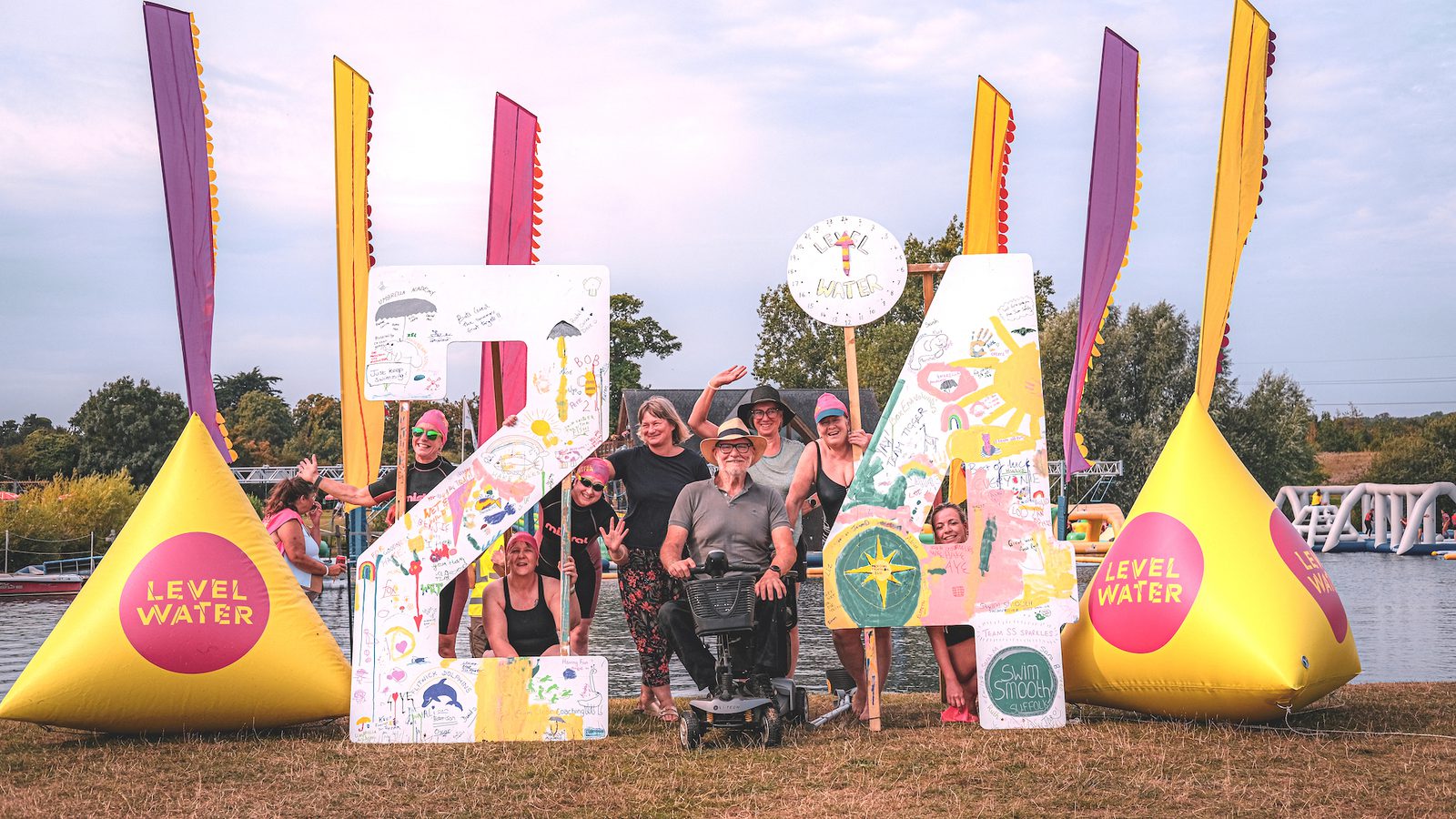 A group of people poses in front of large, colorful numbers "24" and two yellow triangular structures displaying "LEVEL WATER." They appear to be at an outdoor event near a body of water, with trees and inflatables in the background.