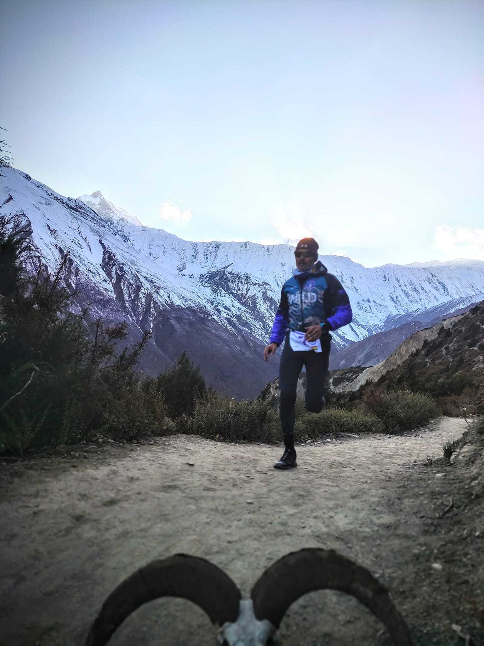 A person is running on a dirt trail surrounded by snow-covered mountains and sparse vegetation. The sky is clear with a hint of light, indicating either morning or evening. The scene exudes a serene, invigorating atmosphere.