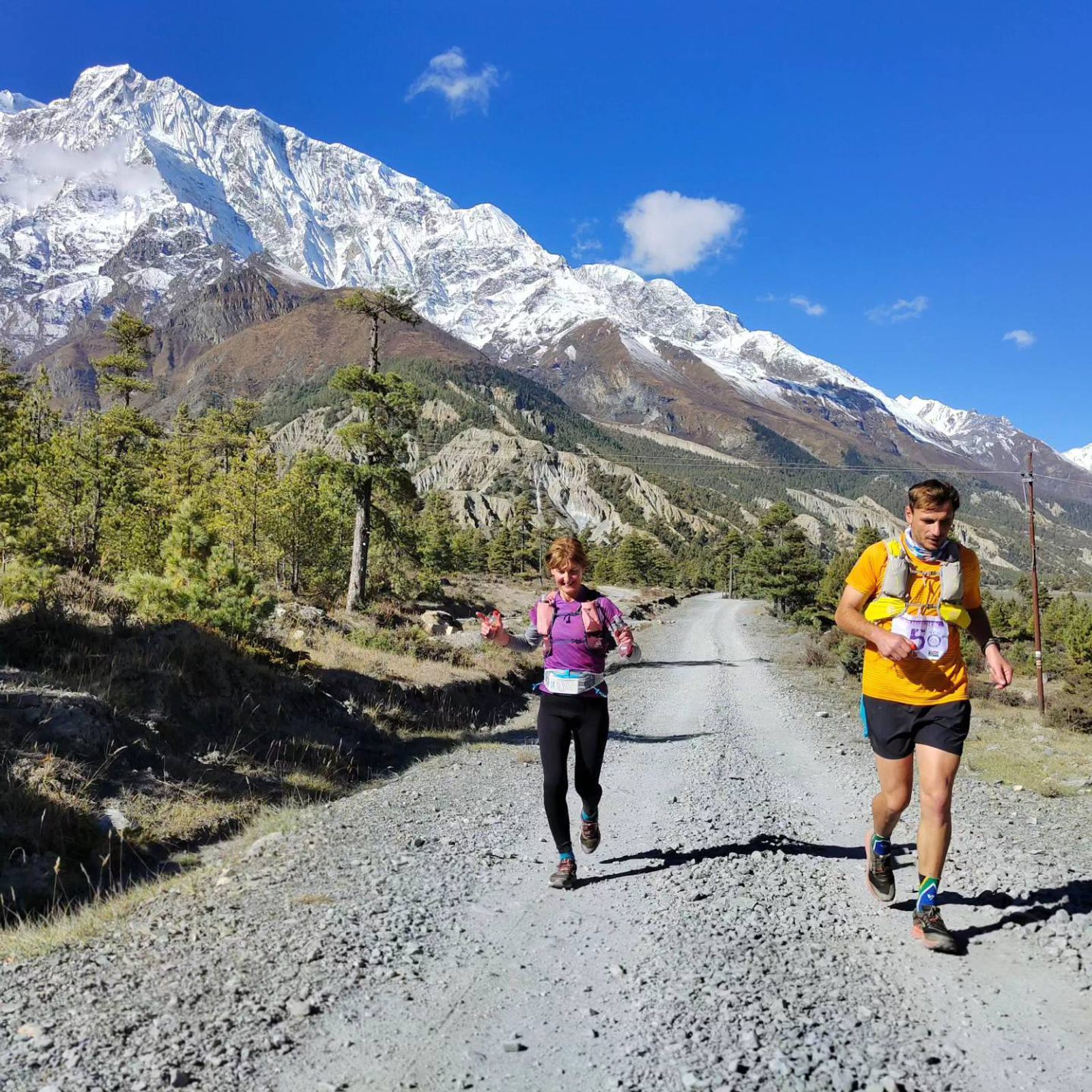 Two runners navigate a gravel path in a mountainous landscape under a clear blue sky. The man on the right wears a yellow shirt and backpack, while the woman on the left sports a purple shirt. Snow-capped mountains and lush greenery form a picturesque backdrop.