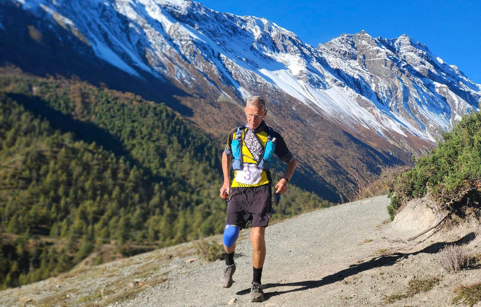 A man is trail running along a mountain path with snow-capped peaks in the background. He is wearing race gear, including a hydration vest and knee support, and is focused on the trail ahead. The scenery includes lush green trees and rugged terrain under a clear blue sky.