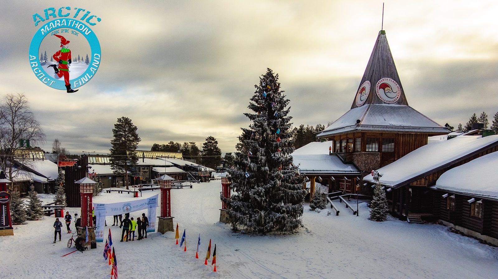 A snow-covered finish line of the Arctic Marathon in Rovaniemi, Finland, marked with flags and a banner. People gather at a large decorated Christmas tree and next to log buildings with a conical tower. The sky is partly cloudy. An Arctic Marathon logo is visible.