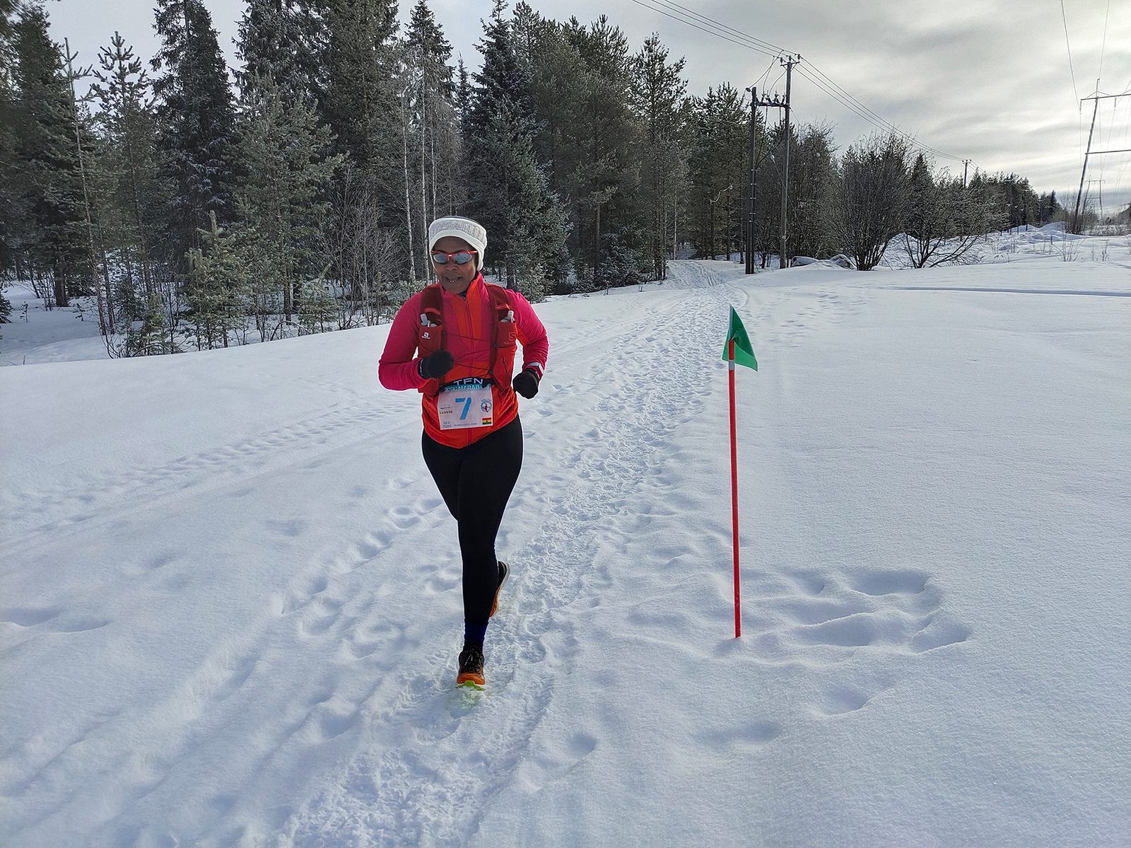 A person wearing a pink top, orange racing bib, black pants, and a white hat runs through a snowy forest trail. They are following a path marked by a red stick with a green flag. Tall evergreen trees and overcast skies can be seen in the background.