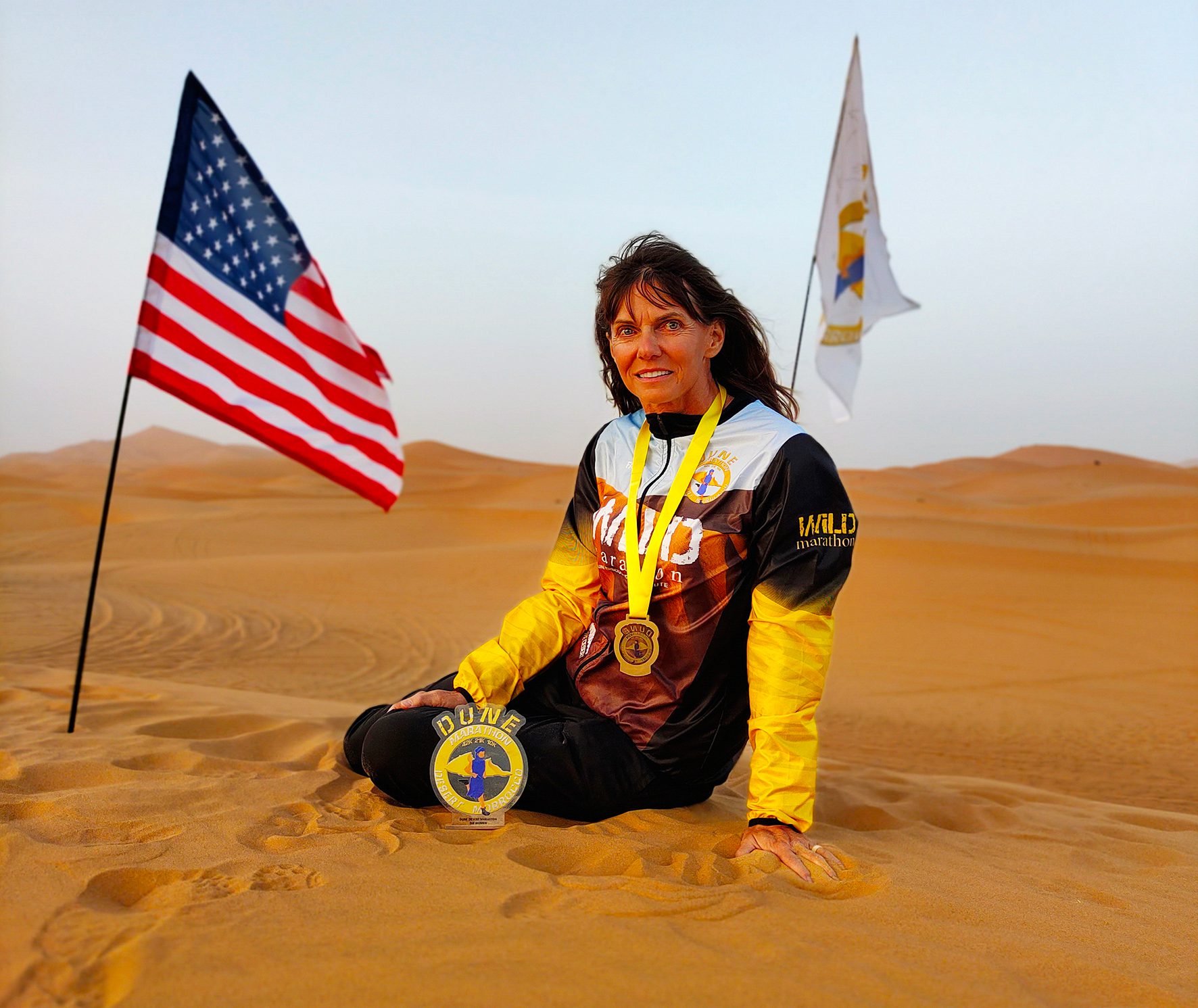 A person with long hair sits on the sand in a desert, wearing a black and yellow sports outfit. They have medals hanging from their neck. An American flag and another flag are planted in the sand behind them. The sky is clear with a warm light.