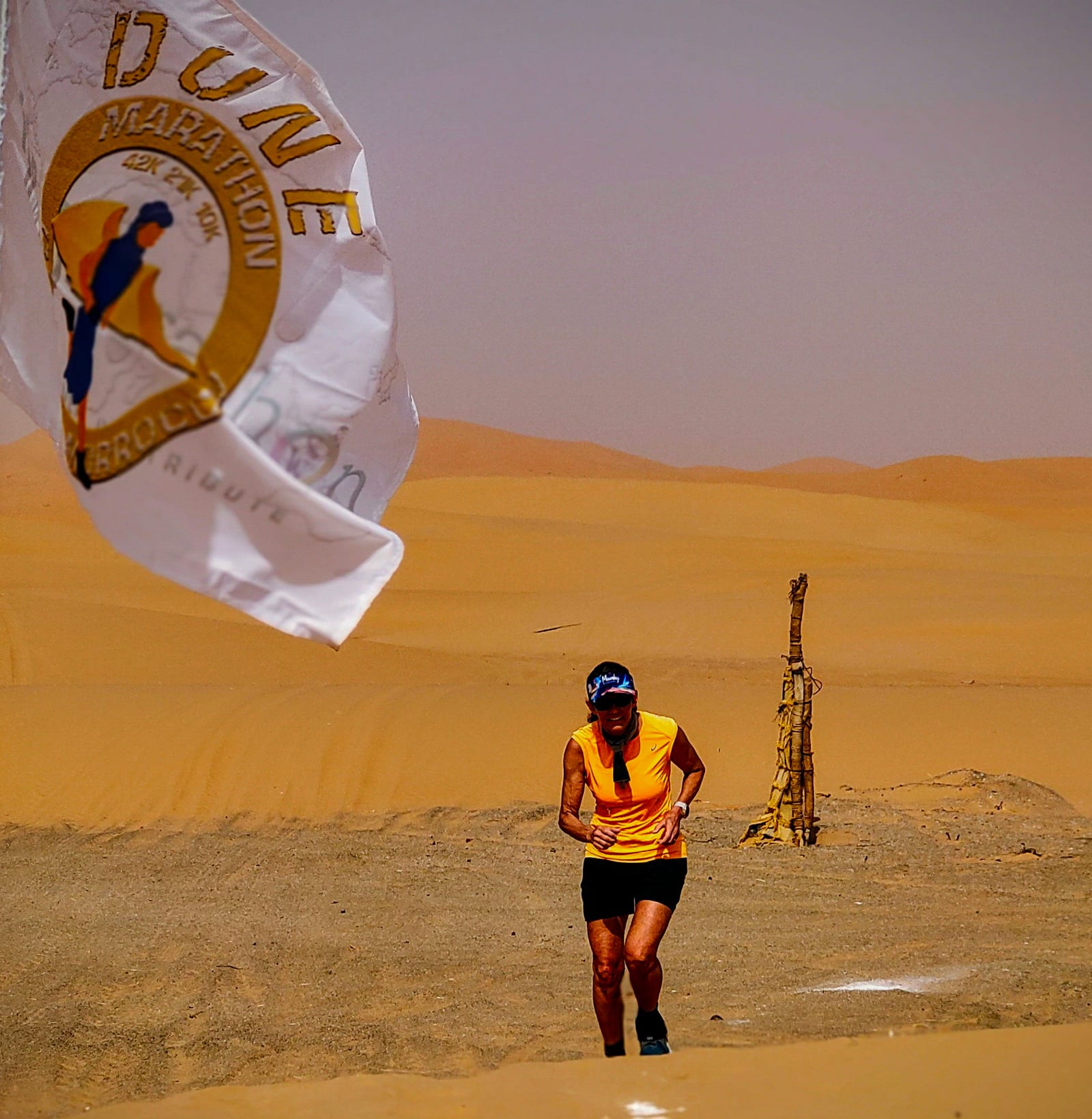 A person wearing an orange shirt, black shorts, and a blue cap is running through the desert with sand dunes in the background. A large white flag with "Dune Marathon" and an image of a runner is prominently displayed on the left side of the image.