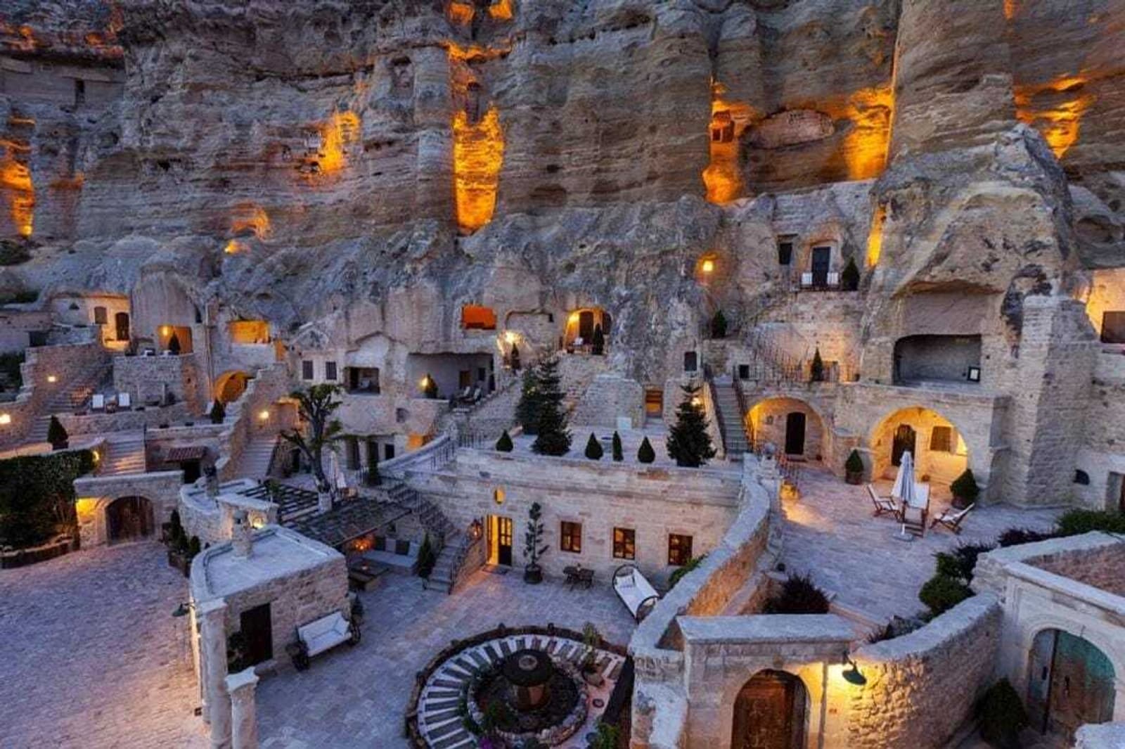 A cliffside hotel with stone buildings and rooms built into the rock, warmly lit at dusk. Courtyards, staircases, and greenery are visible, blending with the rugged rock face above.