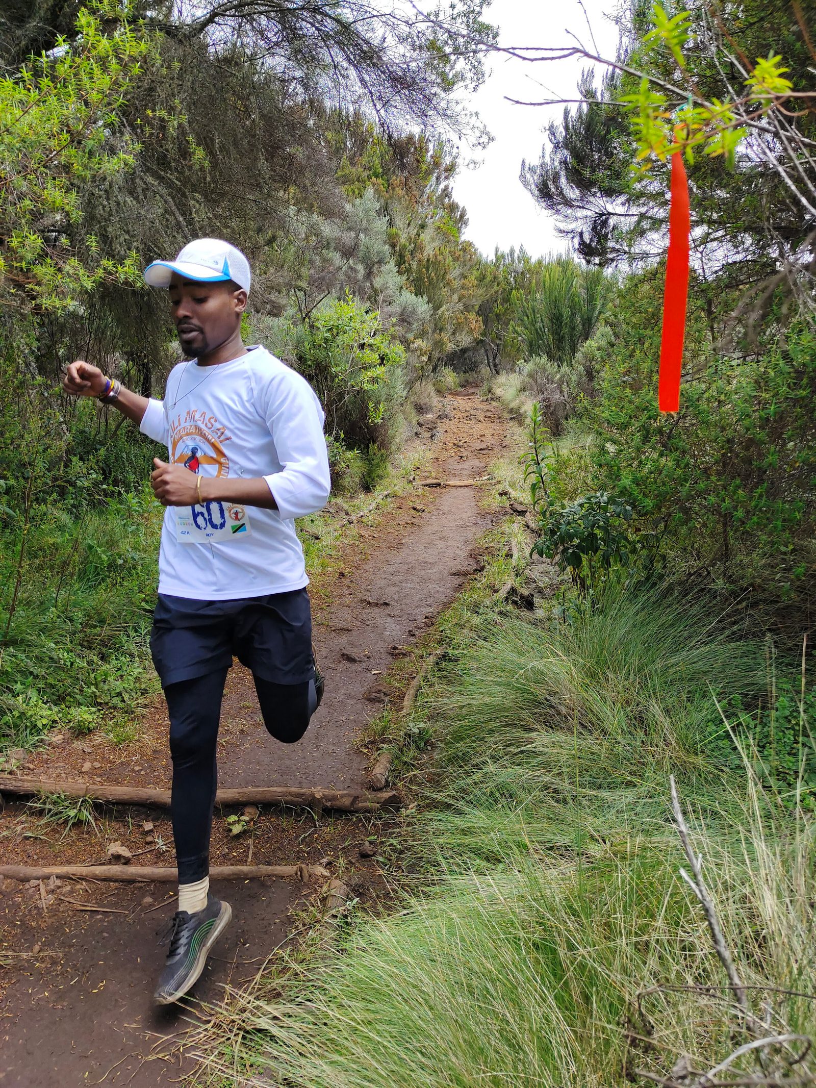 A person wearing a white cap, white long-sleeve shirt, and dark athletic pants is running on a trail surrounded by lush greenery. An orange trail marker hangs from a branch to the right. The runner smiles with an open-mouthed expression as they sprint forward.