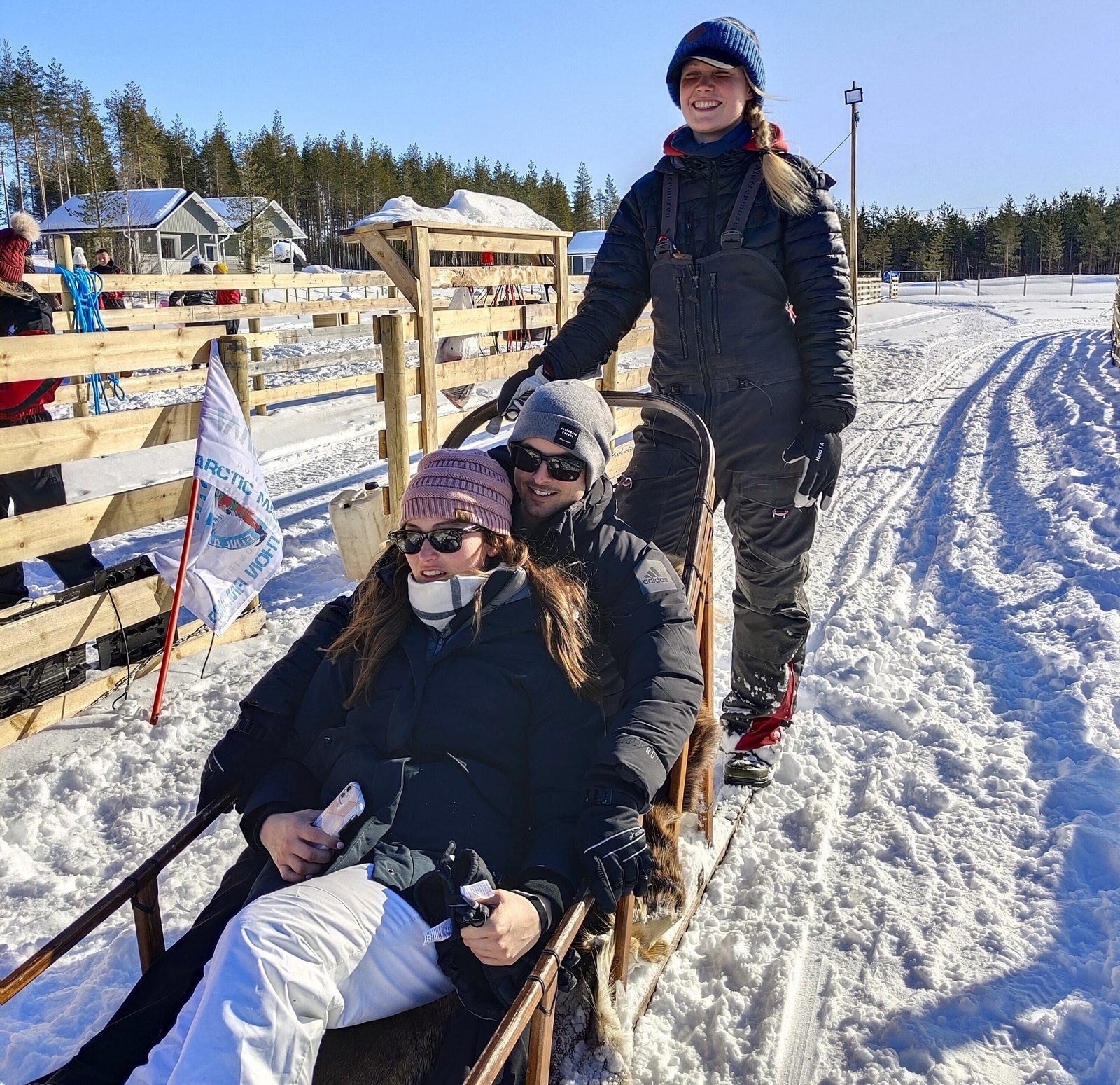 Three people enjoying a dog sledding activity in a snowy landscape. Two individuals are seated in the sled, dressed warmly in winter clothes and sunglasses, while the third stands behind, steering the sled. A wooden fence and other snowy terrain are visible in the background.
