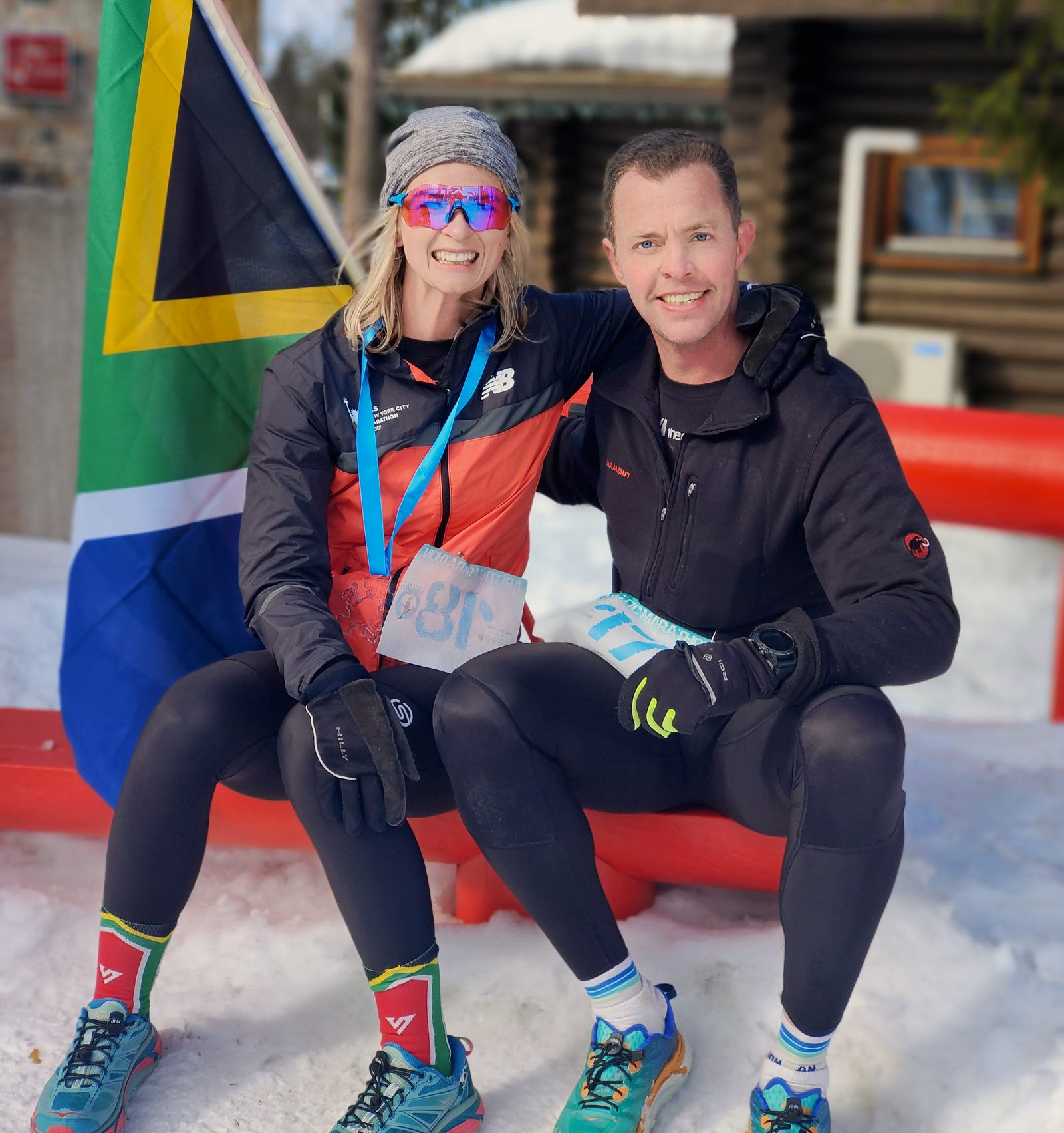 Two smiling runners sit on a red bench in a snowy outdoor setting, with a South African flag in the background. Both are dressed in athletic gear, with the woman wearing sunglasses and a medal around her neck. The man has his arm around her shoulder.