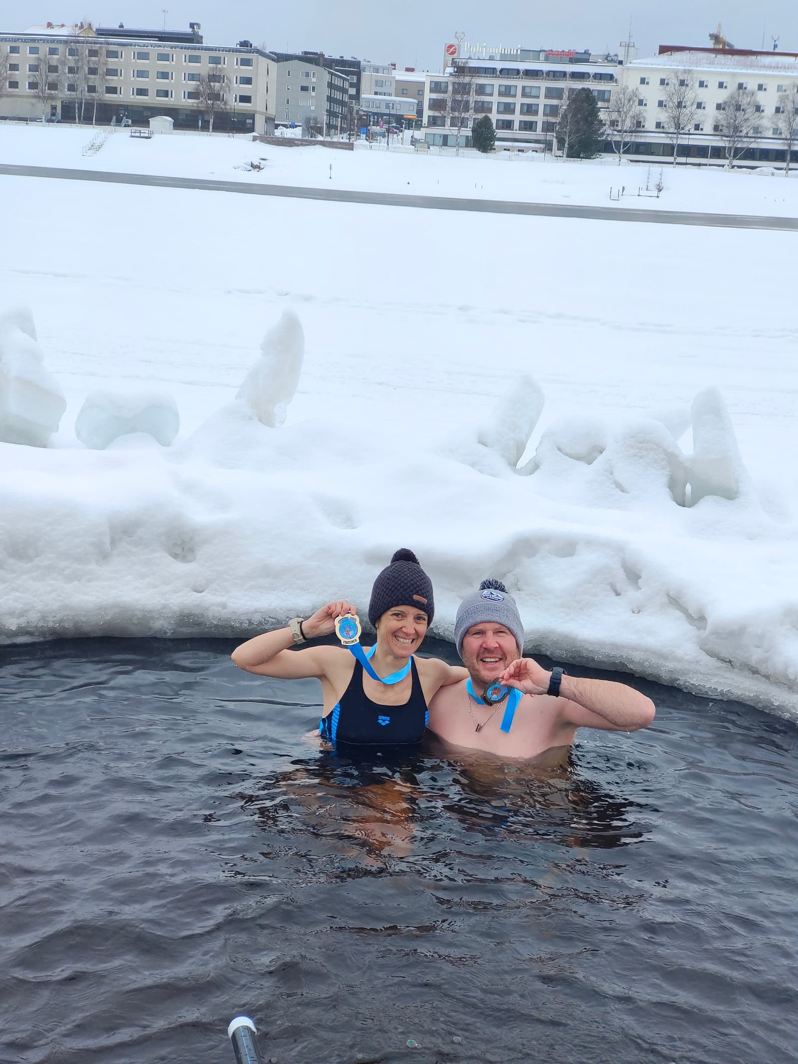 Two people in swimwear and winter hats stand waist-deep in an icy outdoor pool, smiling and holding up medals. Snow and ice surround the pool, with snowy buildings and trees in the background.