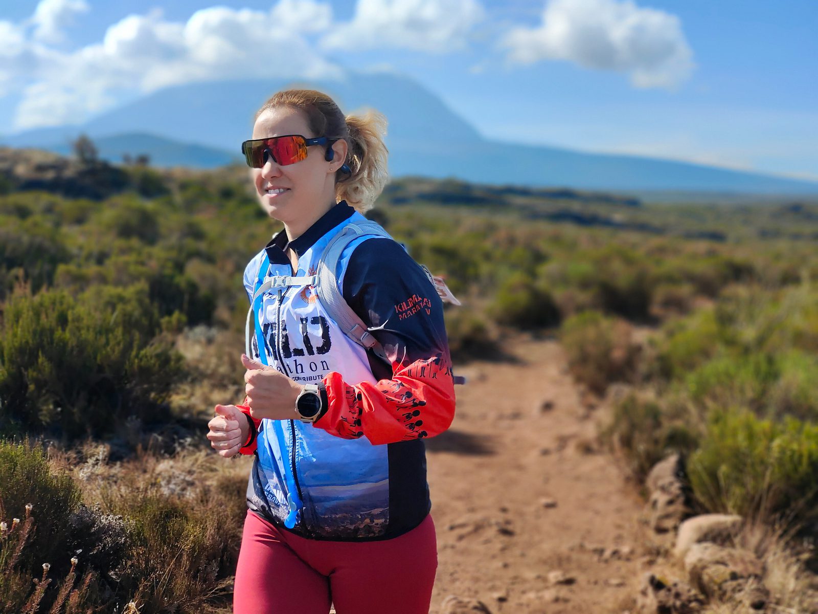 A person with blonde hair wearing sunglasses, a colorful athletic jacket, and red leggings is running on a trail through a scenic, shrub-dotted landscape. The background features a mountain under a bright blue sky with some clouds.