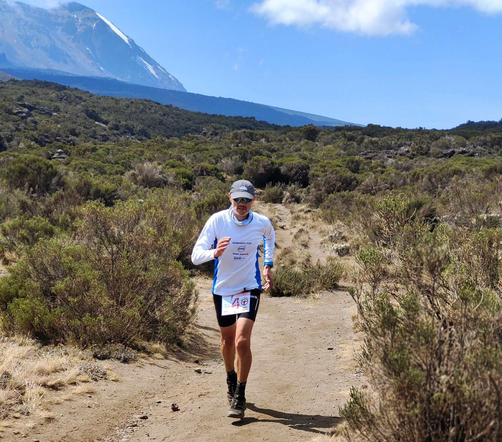 A person wearing a white long-sleeve shirt, shorts, and a visor runs along a dirt trail in a mountainous area. Sparse vegetation and rugged terrain surround the path. In the background, a tall mountain, partially snow-capped, rises under a blue sky with some clouds.