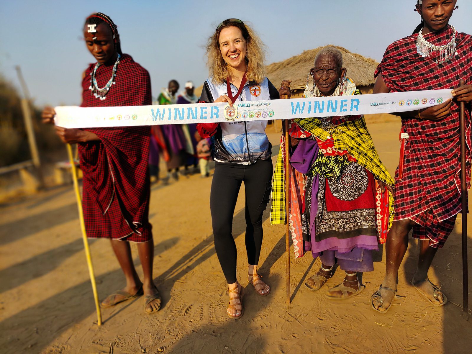 A smiling woman stands holding a "WINNER" banner, flanked by four smiling people in traditional attire. The group is outdoors on a sunny day, with a thatched-roof hut in the background. The atmosphere is celebratory and joyful.