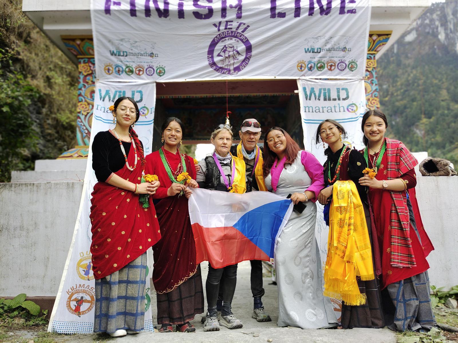 A group of people stands under a "Finish Line" banner. Two people in the center hold a Czech flag, surrounded by others in traditional attire holding flowers. They pose in front of a decorated entrance with mountains in the background.