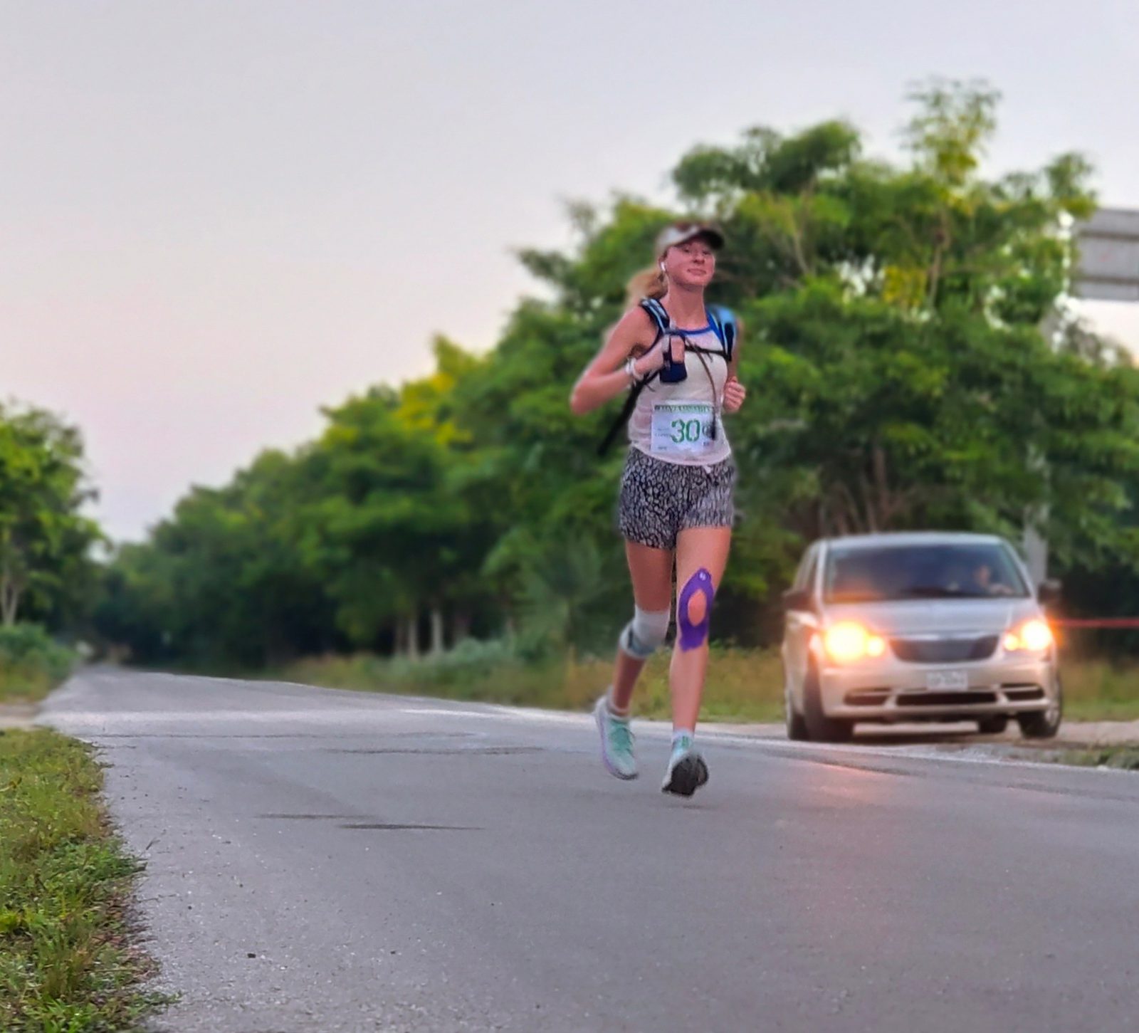 A woman with a "30" race bib is jogging on a paved road with trees in the background. She is wearing a hat, a tank top, patterned shorts, and a knee brace. She is carrying a hydration backpack. A car is driving on the road behind her. The sky is clear.