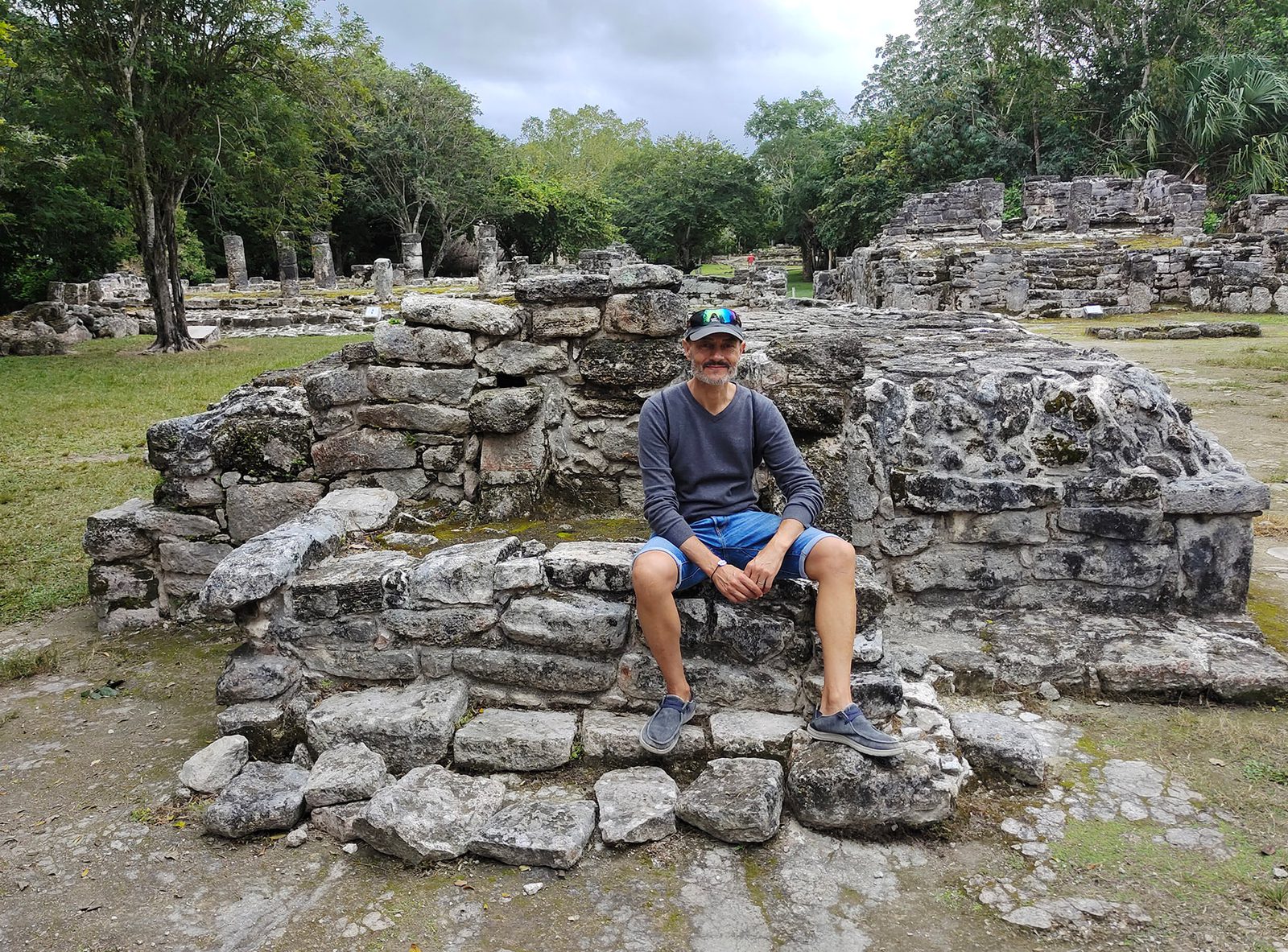 A person wearing a cap, sunglasses, gray shirt, and blue shorts sits on a stone structure in an ancient ruins site surrounded by rough stone walls and lush greenery. The sky is overcast, adding a muted tone to the scene.