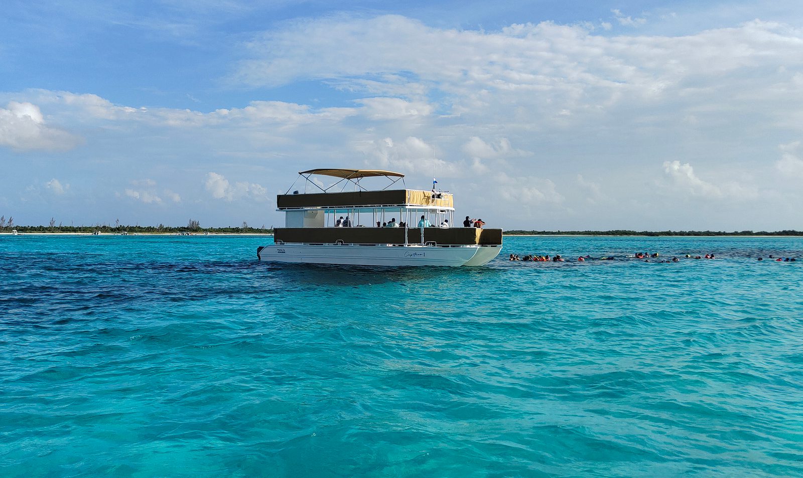A large boat floats on clear turquoise waters under a blue sky with scattered clouds. Near the boat, a group of people can be seen swimming and snorkeling, forming a line parallel to the boat. A distant shoreline is visible in the background.