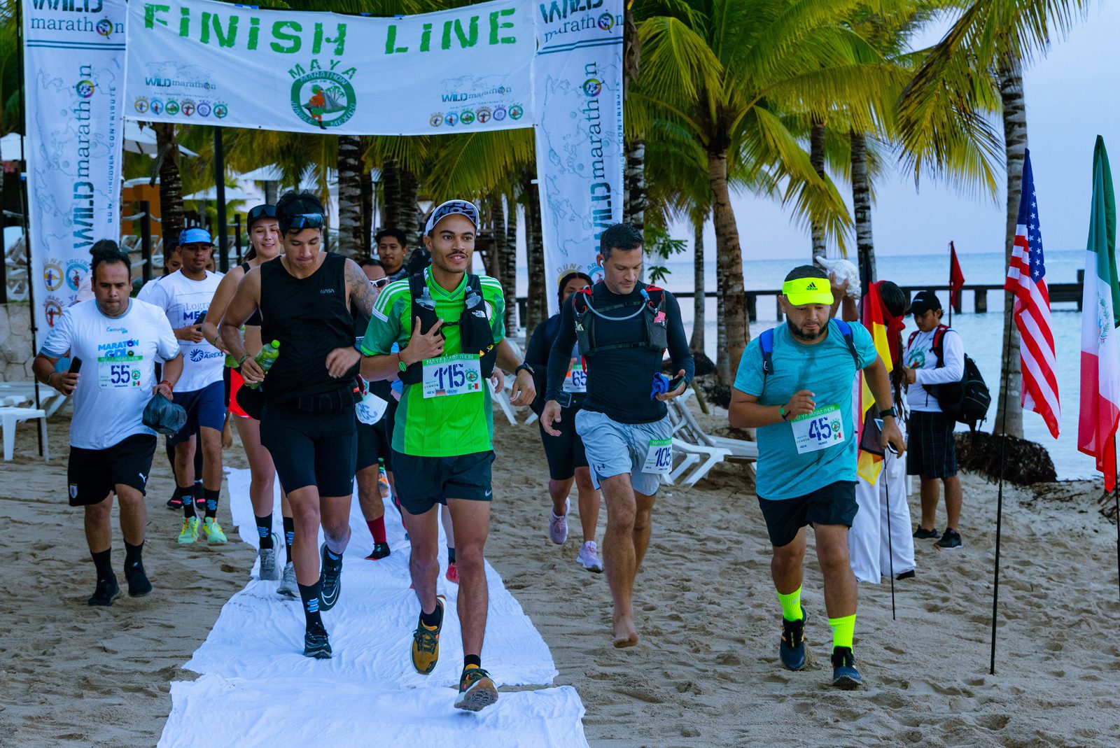 Runners cross a finish line on a beach, flanked by palm trees. Several athletes, wearing bib numbers and athletic gear, jog on a white carpet. An international array of flags, including the USA and Mexico, is visible in the background. Signs read "Finish Line" and "Wild Marathon.