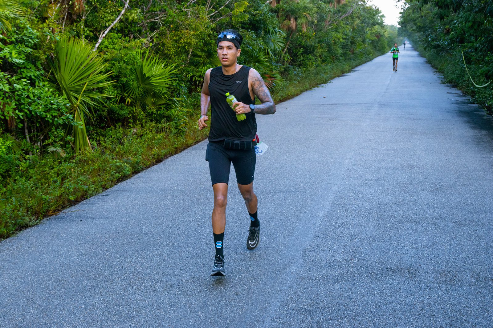 A person wearing black athletic gear and a headband is running on a paved path through a lush green forest. A water bottle is in their right hand. Another runner is seen in the distance.