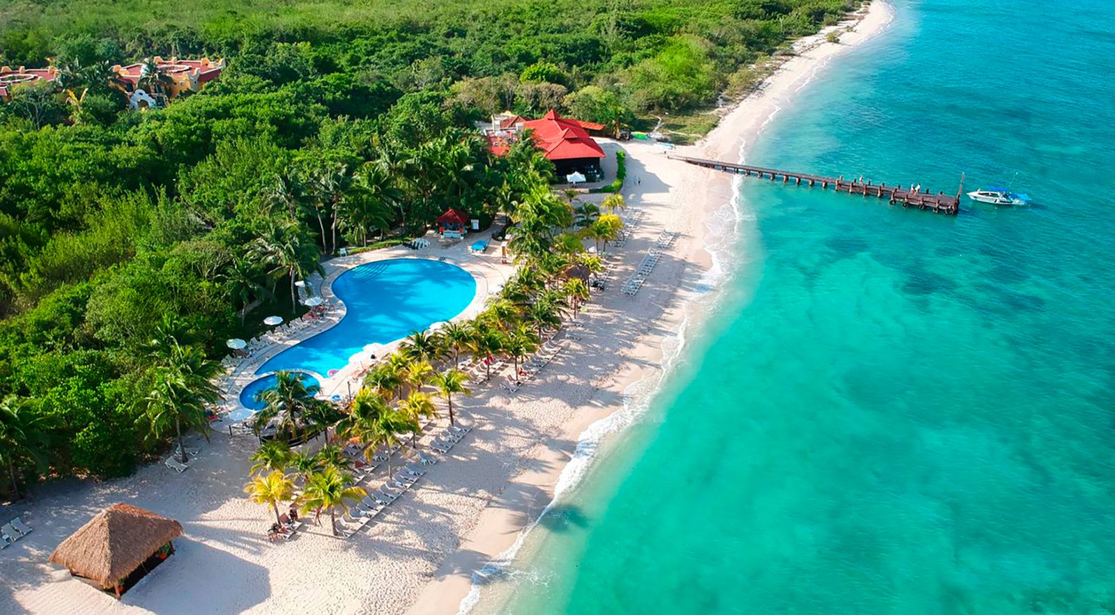 Aerial view of a tropical beach resort with a large, blue swimming pool surrounded by palm trees. The sandy beach stretches along turquoise waters, punctuated by a wooden pier extending into the sea. Lush green vegetation surrounds the resort area.