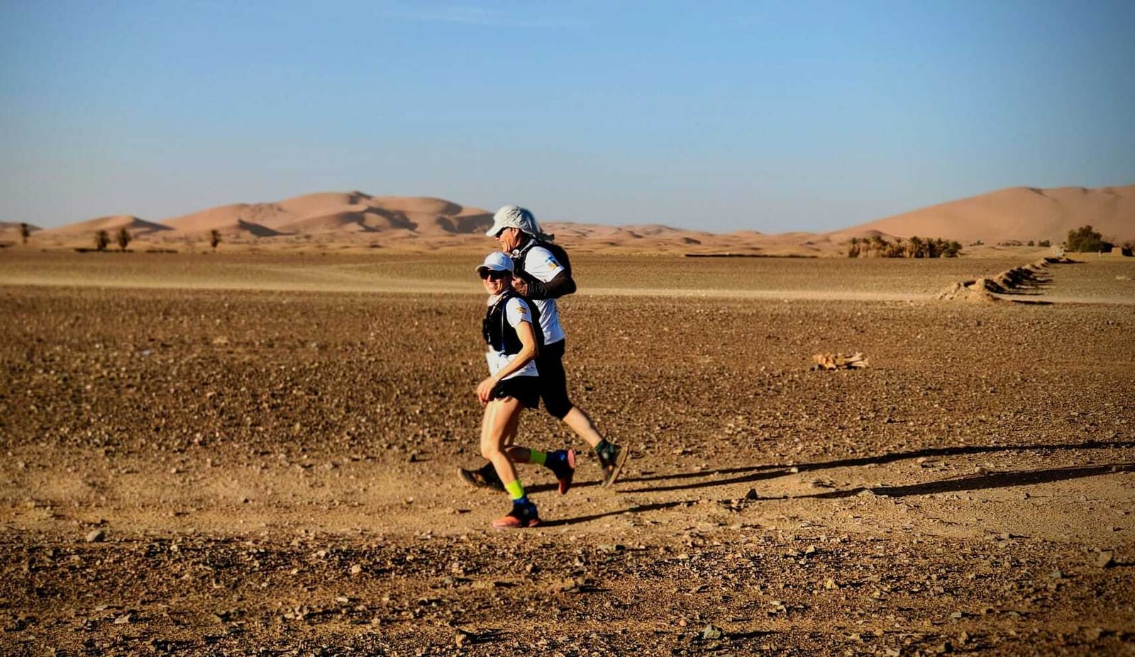 Two people running on a rocky, sandy trail in a desert landscape. They are wearing sunglasses, hats, and backpacks under a clear blue sky, with sand dunes visible in the background.