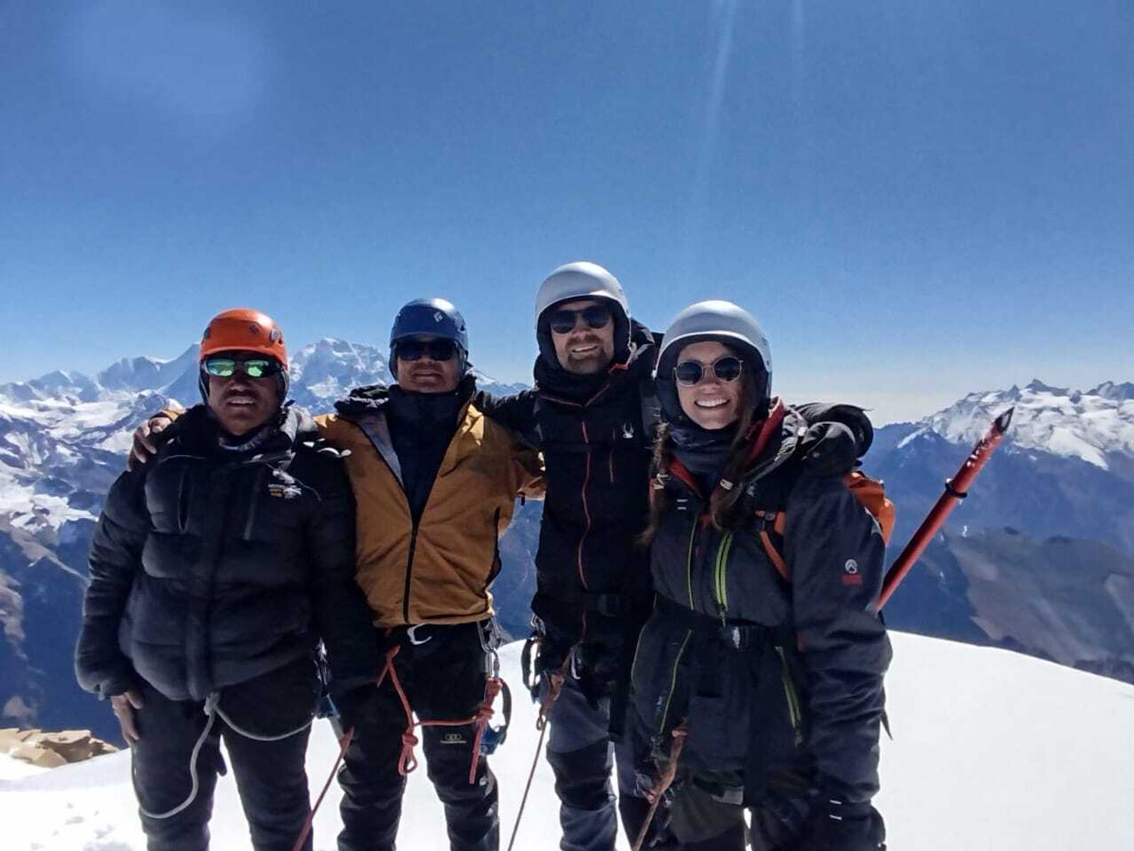 Four climbers in winter gear stand on a snow-covered mountain peak under a clear blue sky, with snow-capped mountains in the background. They are smiling and have their arms around each other.