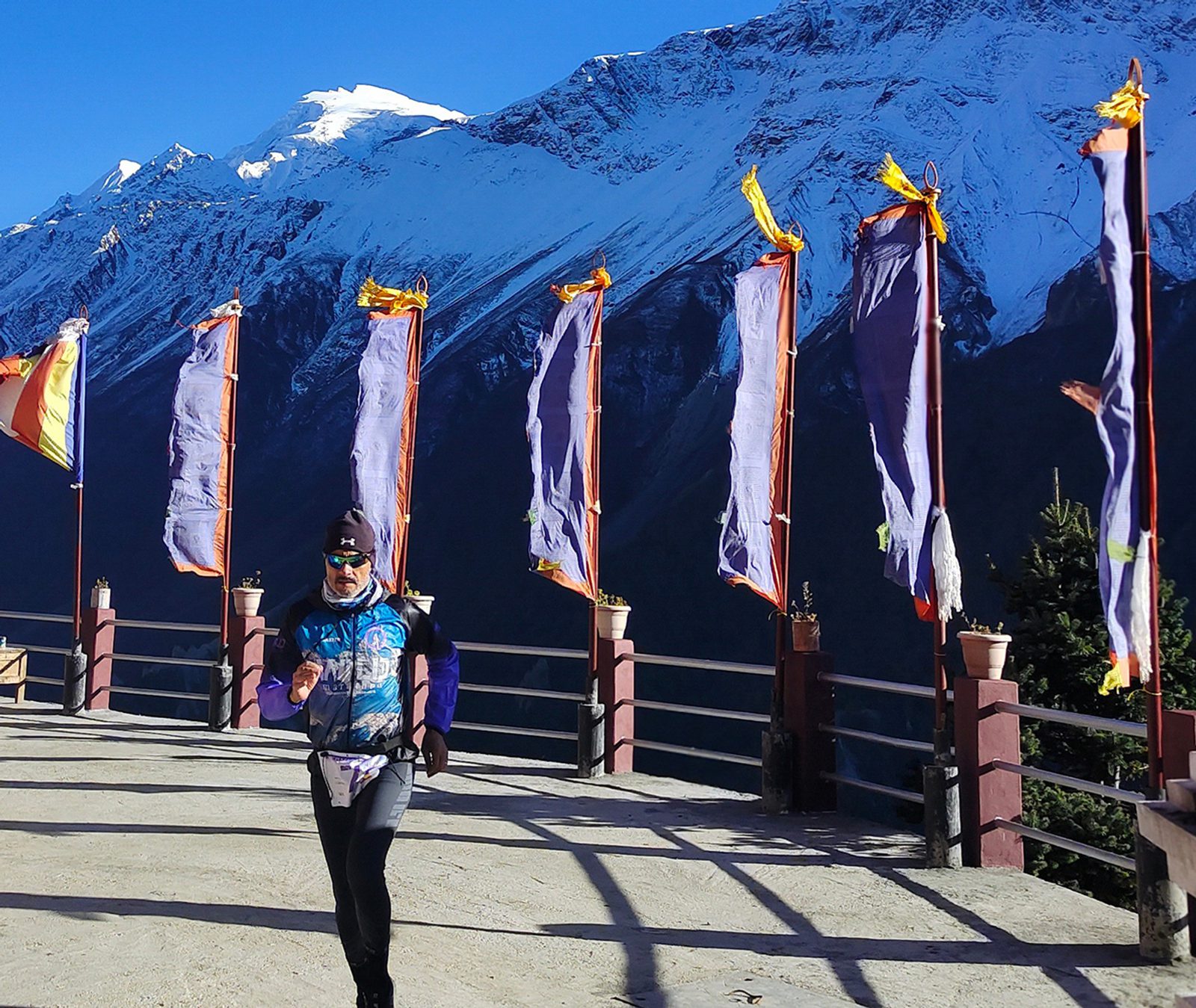 A person wearing outdoor gear runs along a path lined with tall, colorful flags. The backdrop features a stunning mountain range topped with snow under a clear blue sky. The sun casts shadows on the ground. The flags flap in the wind, adding a dynamic element to the scene.