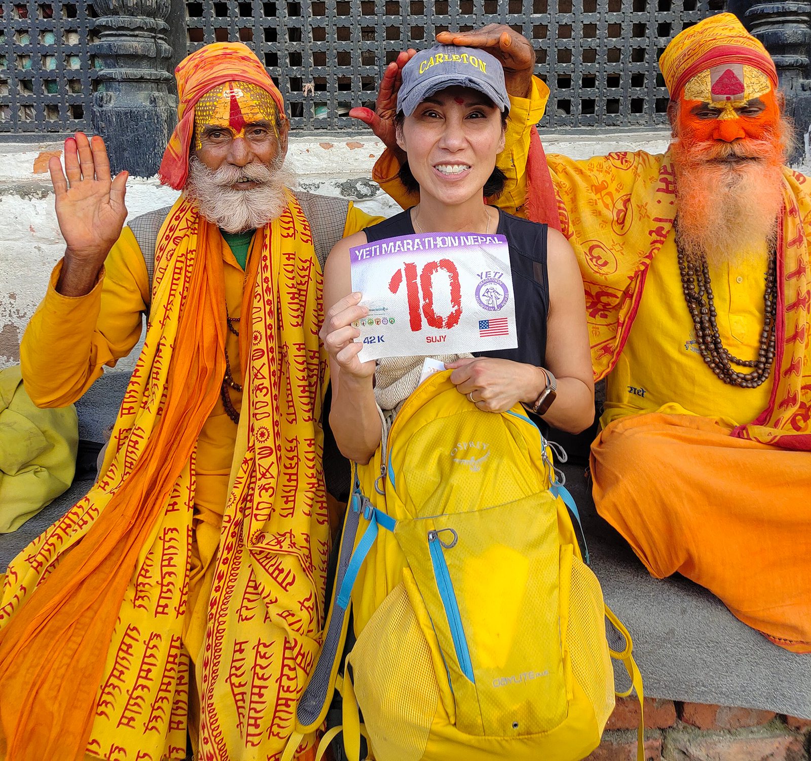 A smiling woman wearing a marathon cap holds a race bib while sitting between two men dressed in traditional orange attire with painted faces. One man raises his hand in greeting, and the other man poses with a cheerful expression.