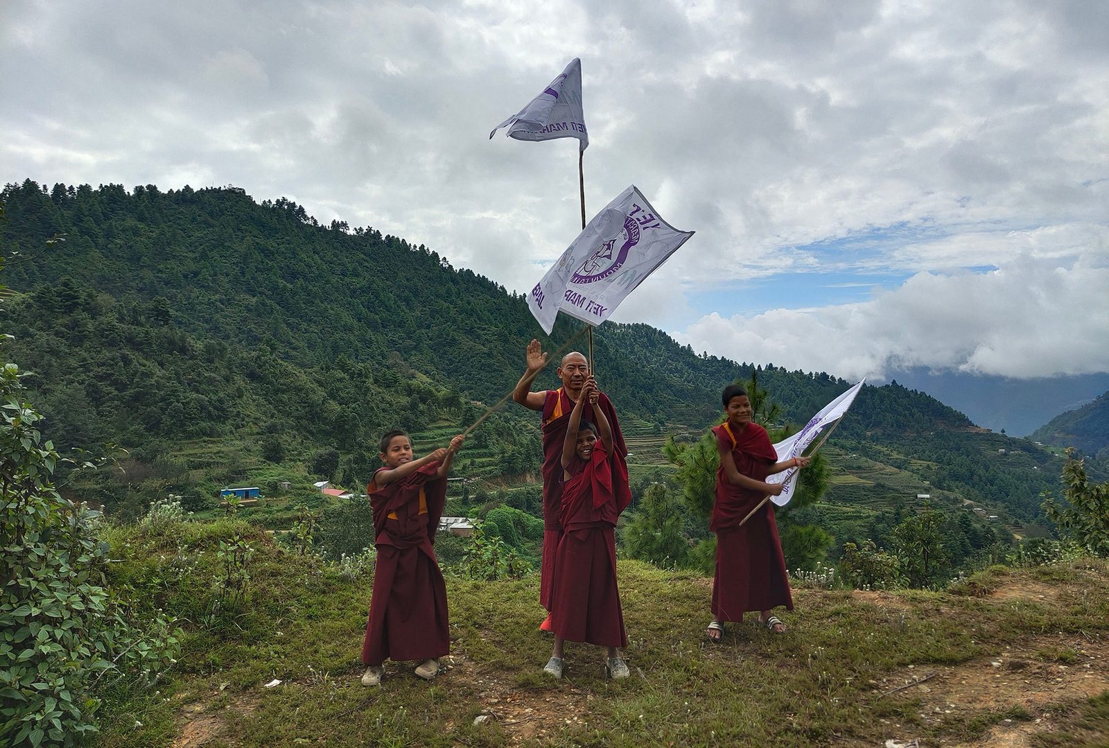 A group of four people in traditional clothing stand on a hillside with lush greenery and mountains in the background. They are holding white flags with a purple emblem, smiling, and appear to be celebrating. The sky is cloudy.