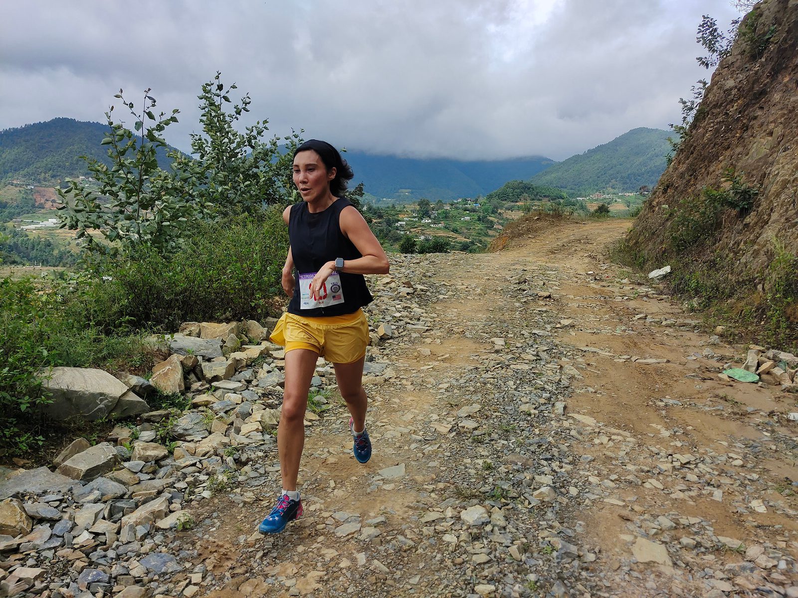 A person is running on a rocky, uneven trail through a hilly landscape. They are wearing a black tank top, yellow shorts, and athletic shoes. The sky is cloudy, and green hills are visible in the background. Some foliage lines the path.