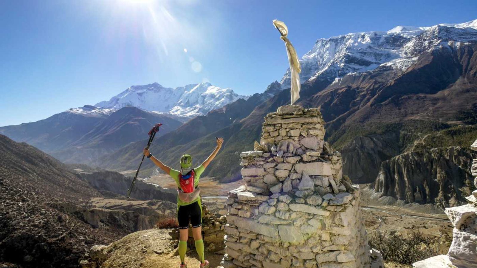 A hiker in green attire and a red backpack raises their arms triumphantly near a stone cairn and prayer flags on a mountain trail. Snow-capped peaks are in the background under a clear blue sky with the sun shining brightly. The landscape is rugged and expansive.