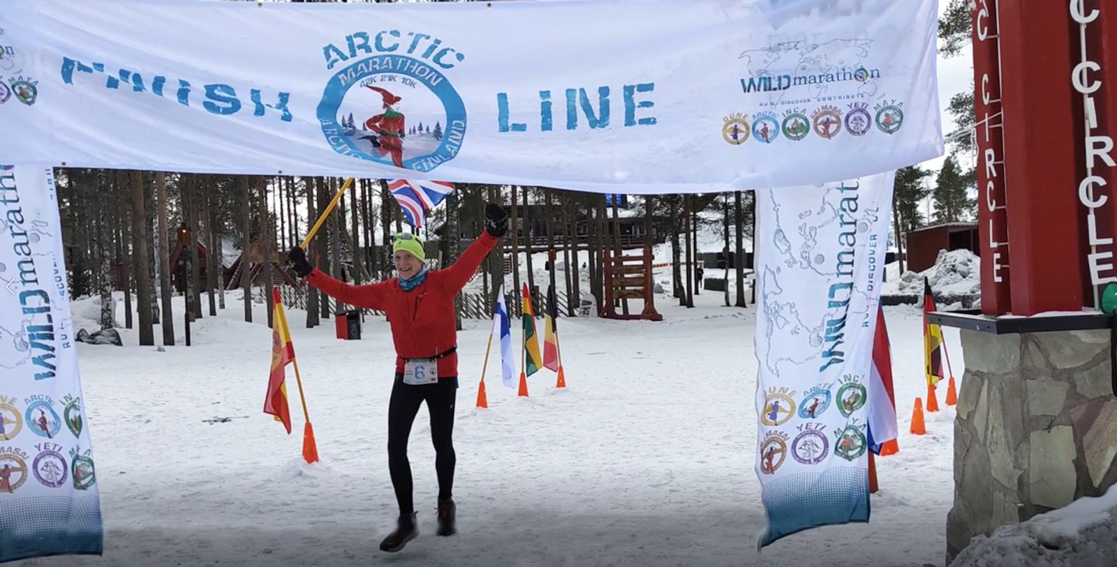 A person wearing winter gear and holding small flags crosses the finish line at an Arctic Marathon event. The snow-covered ground and trees in the background indicate a cold climate, while banners and a red post mark the finish line.