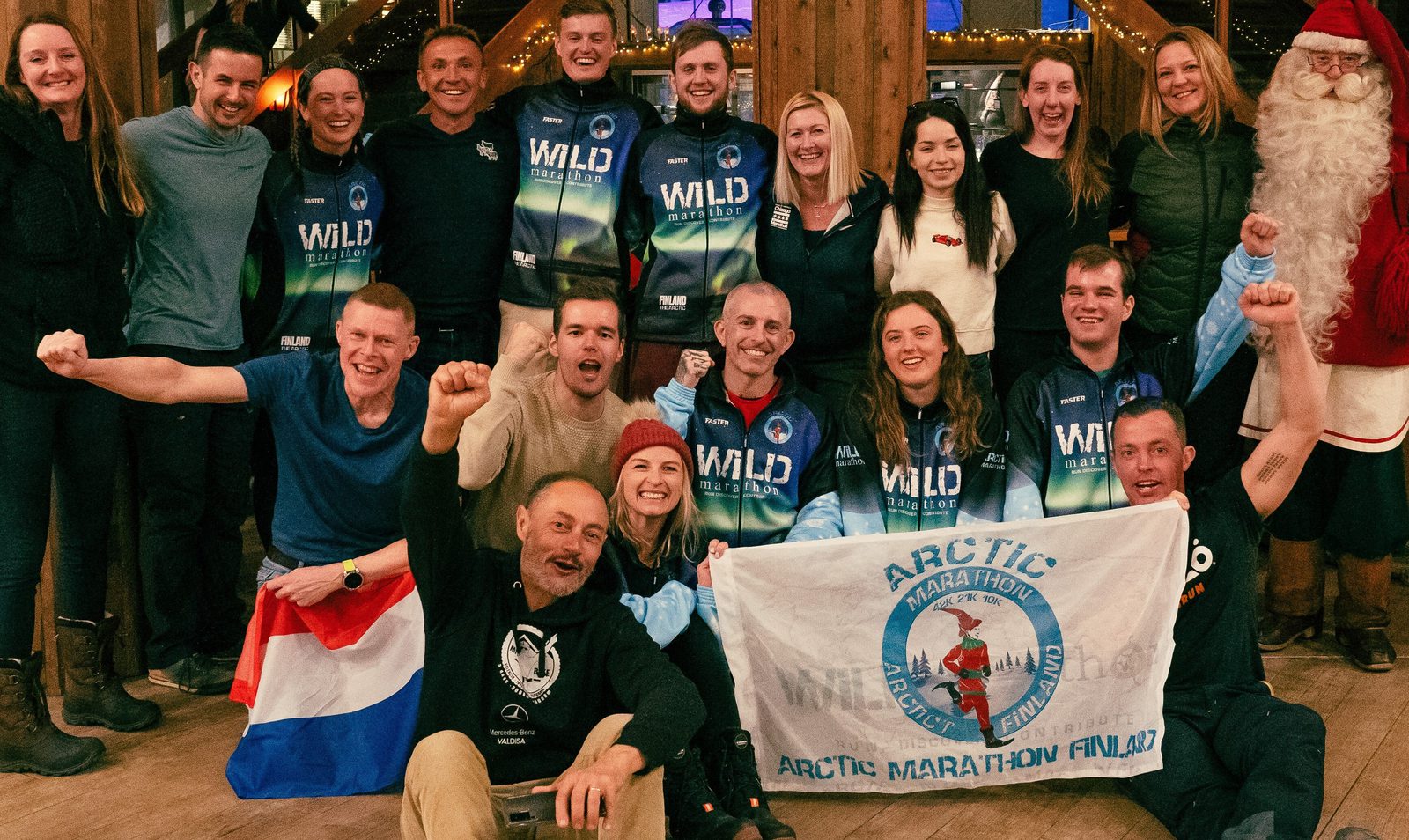 A group of smiling people posing together indoors. Some are wearing matching "Wild" jackets, while others are casually dressed. They are holding a flag that reads "Arctic Marathon, Finland." A person dressed as Santa Claus stands to the right. The atmosphere is festive and celebratory.