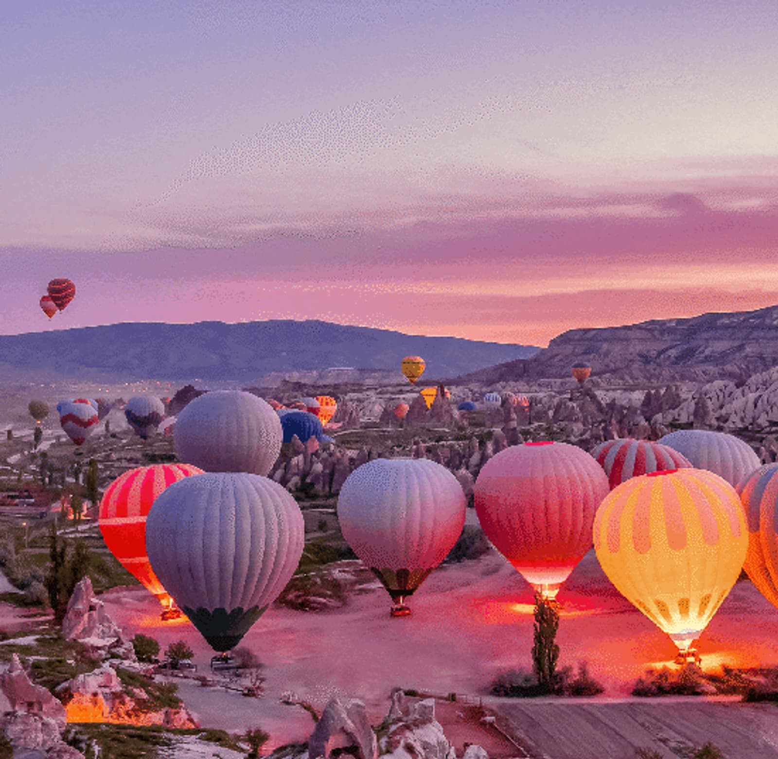 Colorful hot air balloons prepare for takeoff at sunrise in a rocky, mountainous landscape, with a pink and purple sky in the background.