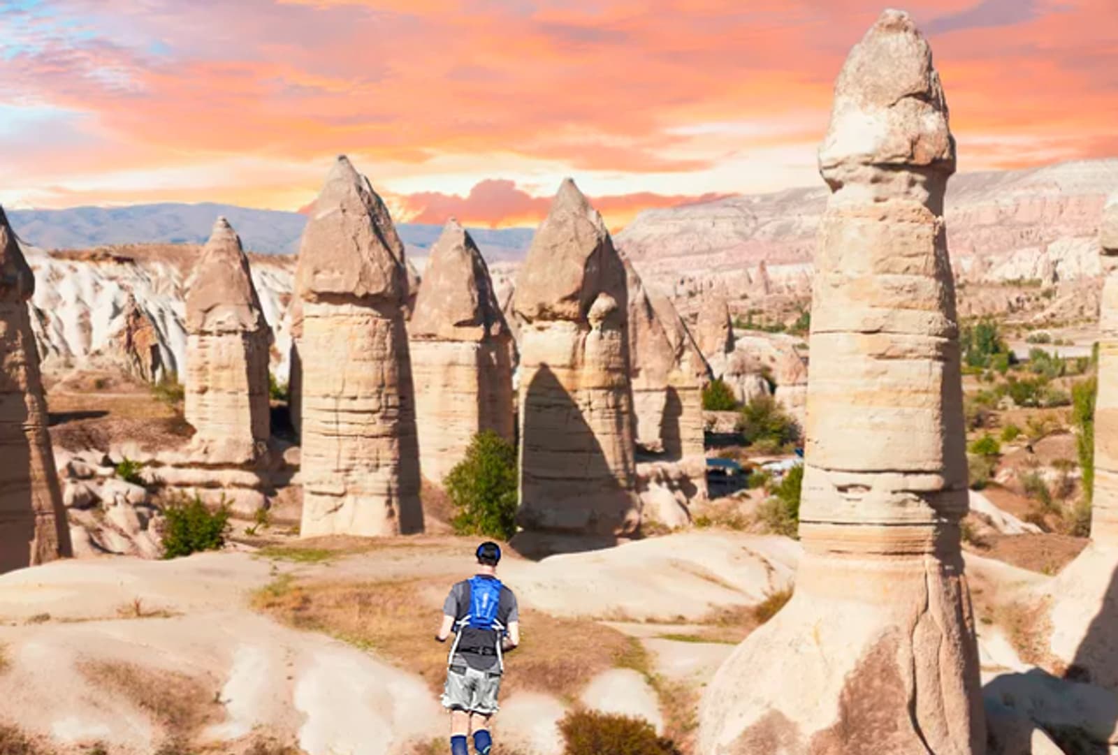A person wearing a blue shirt and cap stands on rocky terrain, facing tall, uniquely shaped rock formations under a colorful sunset sky in Cappadocia, Turkey.
