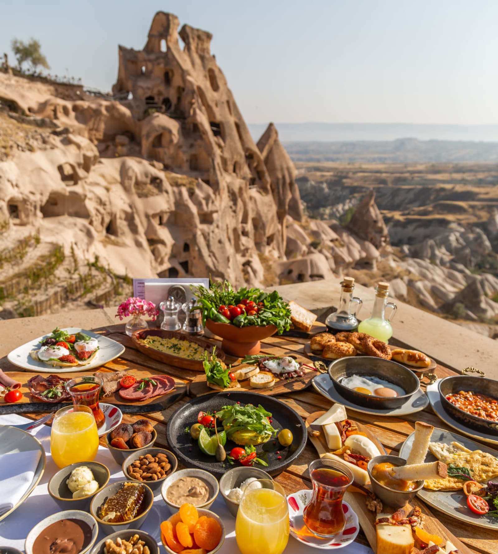 A table set with a variety of colorful Turkish breakfast dishes overlooks unique rock formations and a scenic valley, likely in Cappadocia, Turkey, under a clear sky.