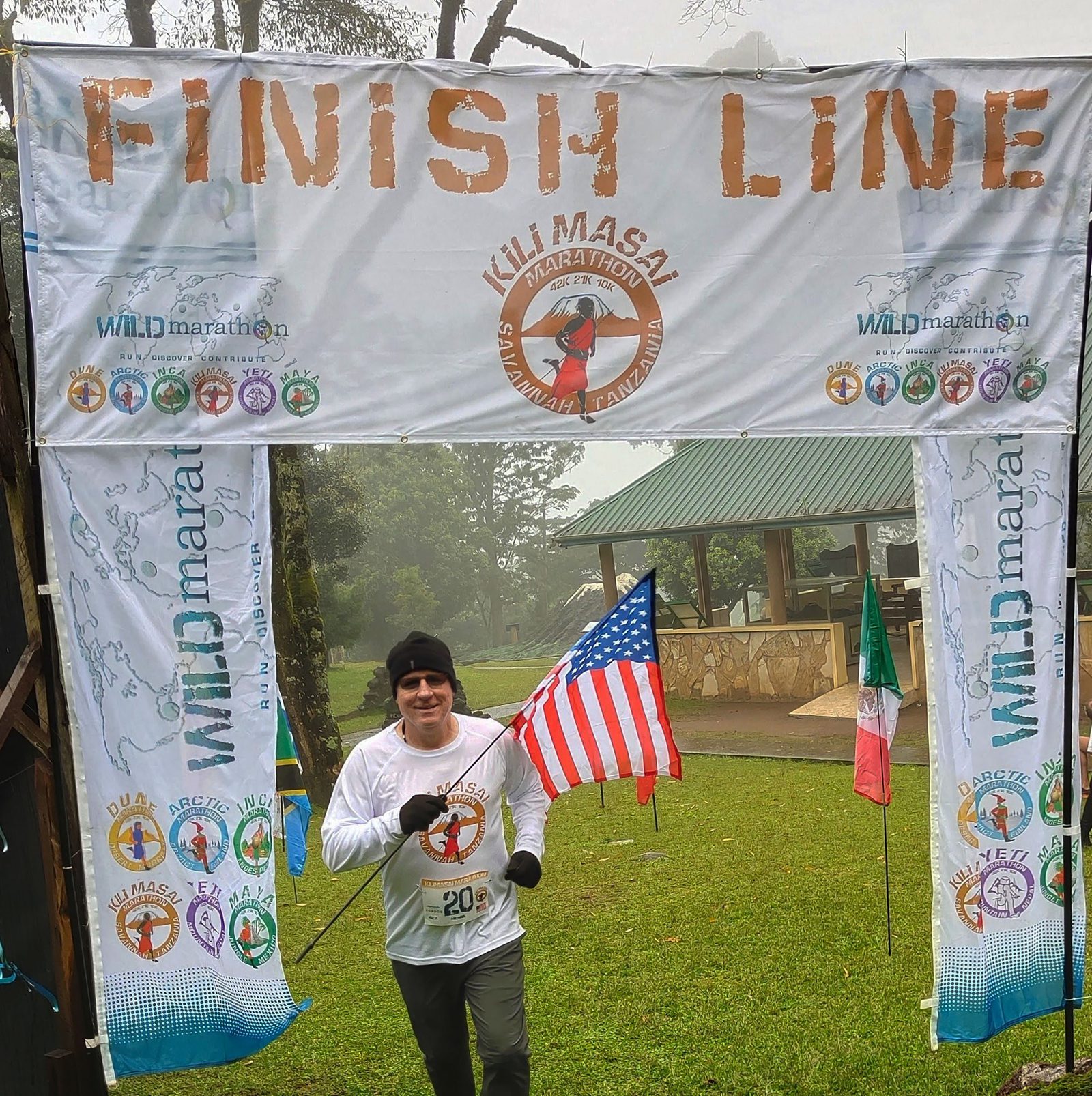 A runner crosses the finish line at the "WILD marathon" event, held outdoors with a foggy background. The runner, wearing a white shirt and black cap, is holding an American flag. The finish line is marked with banners showcasing the event's logo and sponsors.
