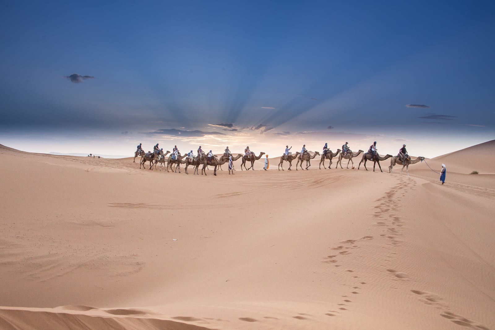 A caravan of camels being led through a vast desert with a person in blue clothing walking beside them. The landscape is covered in sand dunes, and the sun is setting in the sky, casting rays of light through the clouds.