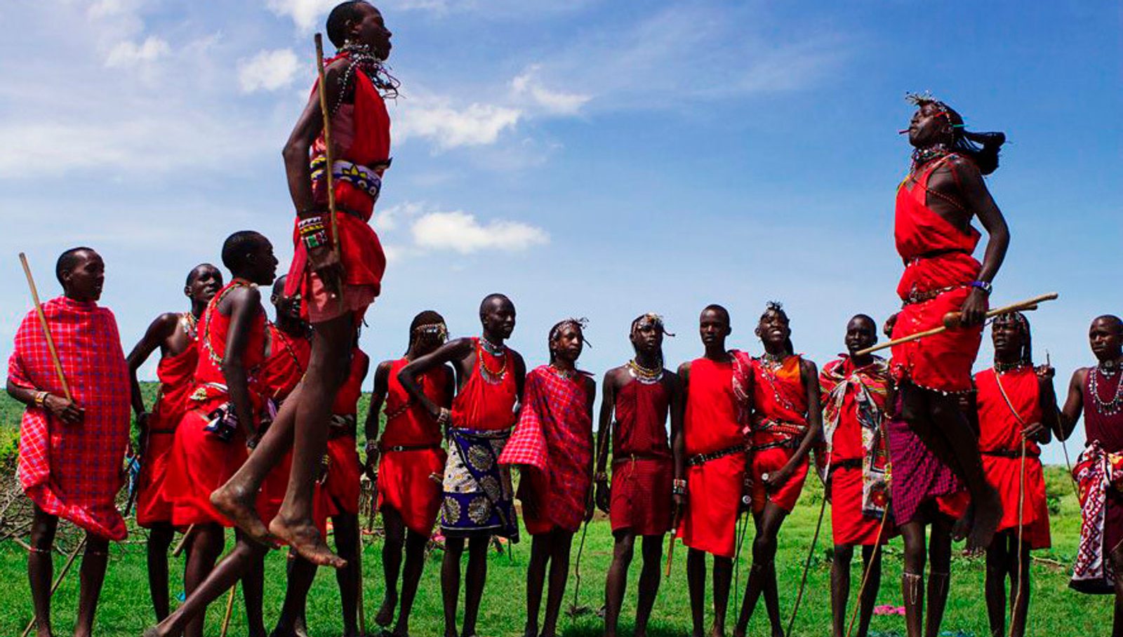 A group of people dressed in traditional red garments perform a dance outdoors. Some are in mid-air, leaping with impressive height, while others form a circle, observing and participating. The background is a clear blue sky with a few clouds.