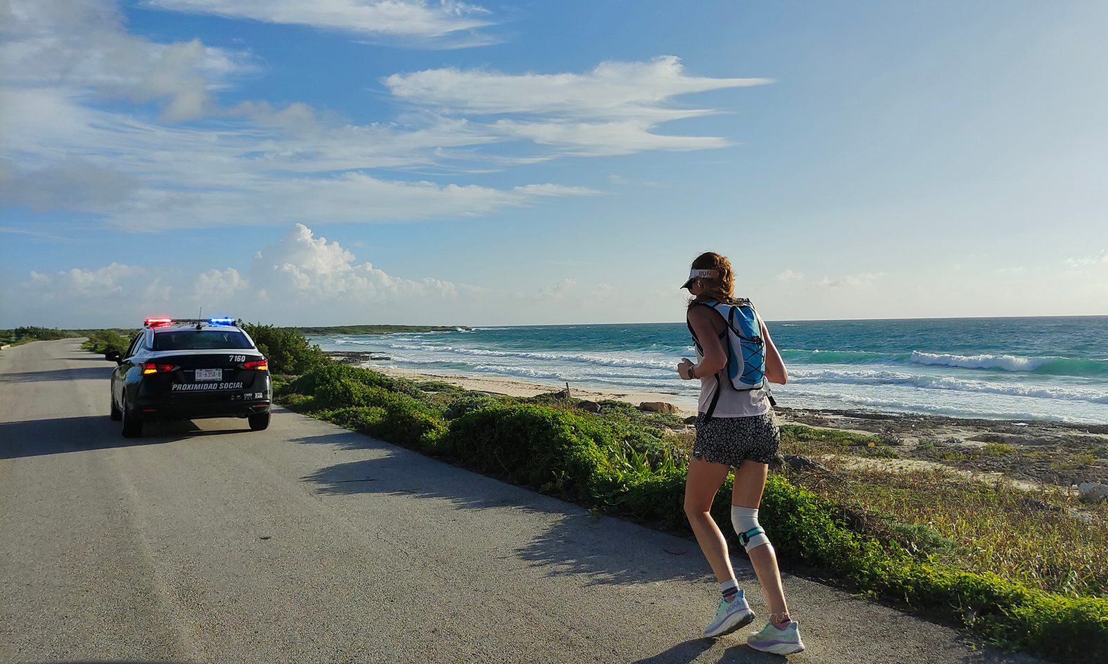 A runner wearing a hat, tank top, and shorts jogs along a coastal road with lush greenery on one side and the ocean on the other. A police vehicle with flashing lights follows on the same road under a clear blue sky with fluffy clouds.